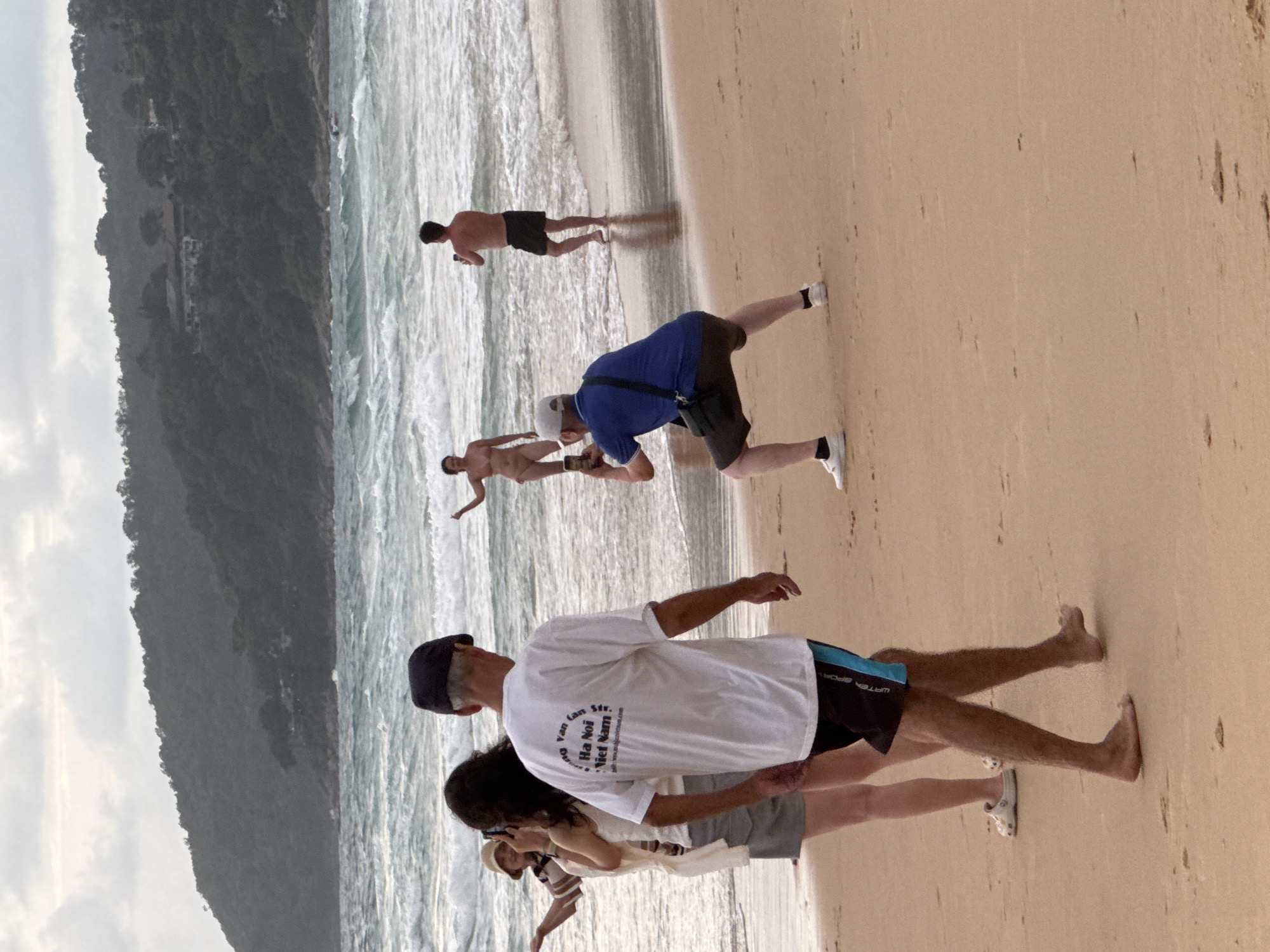 The image depicts a beach scene in Karon, Thailand, with the ocean and a tree-covered hill in the background. Several individuals are present on the sandy shore. In the foreground, a person wearing a white t-shirt and shorts walks away from the camera. To their left, a woman in a striped top and shorts is taking a picture with her phone. Further back, a person in a blue shirt and shorts is kneeling and taking a photo with a smartphone. In the water, a partially visible person in black shorts is holding a phone. To the far left, a woman in a tan hat and striped shirt is standing near the water's edge. In the middle ground, a nude individual is standing in the shallow water. The sky is overcast.