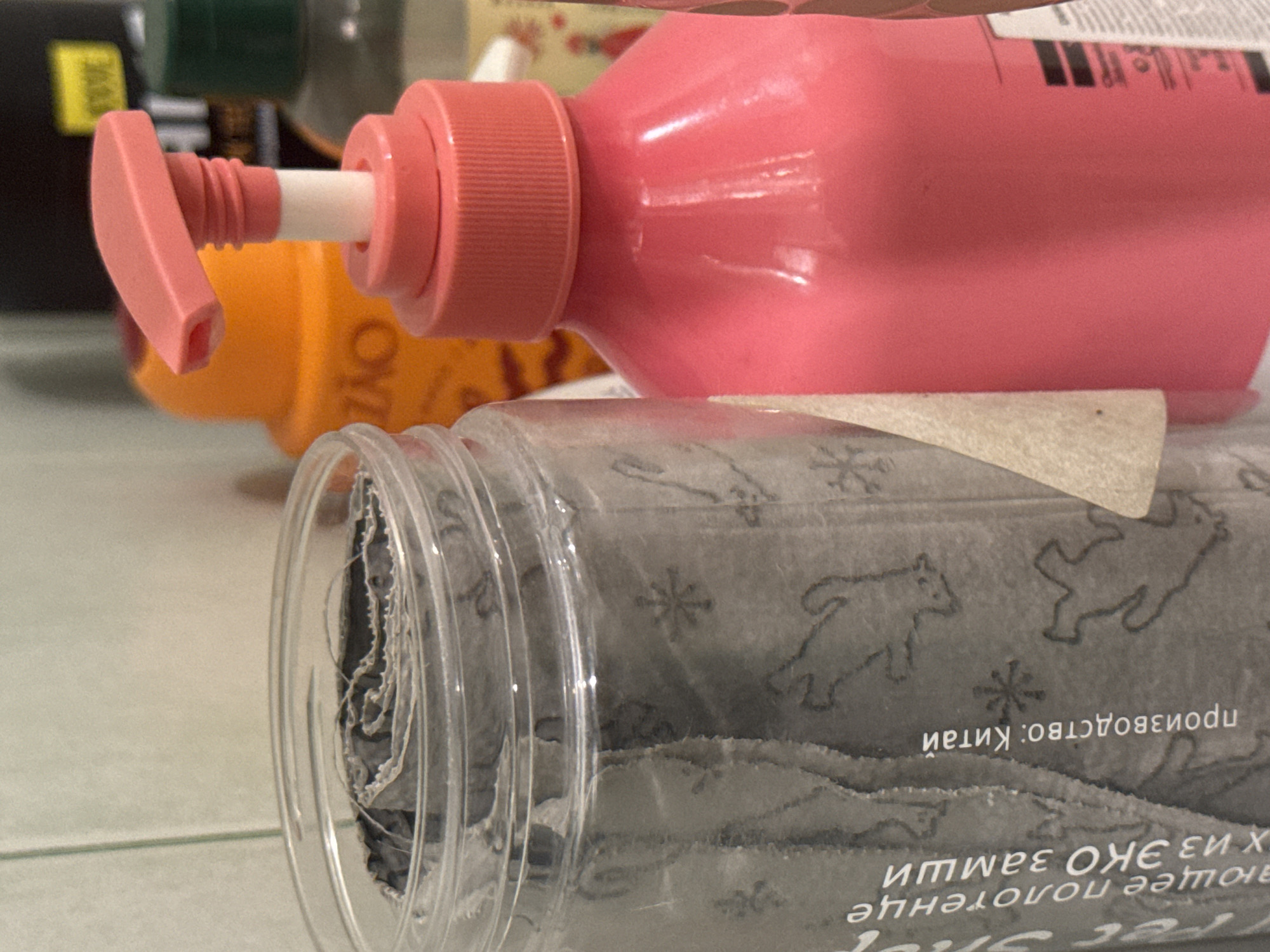 The image shows a close-up of a pink pump dispenser bottle and a clear plastic container with rolled material inside. The pink dispenser is positioned diagonally across the frame, with its pump mechanism visible on the left. The dispenser has a textured pink cap and a white tube extending from it. In the background, out of focus, are other bottles, one of which appears to be orange and has some text on it.
Below the pink dispenser, and slightly out of focus, is a clear plastic container. The container's ribbed texture is visible along its edge. Inside, a roll of dark gray material is visible. The material has a repeating pattern of silhouetted animals, possibly dogs or wolves, and small star-like shapes. Some text is visible on the rolled material, appearing to be in Russian. One line of text is oriented upside down and reads "производство: Китай" (production: China). Another line of text, also upside down, reads "из эко полотенце" (from eco towel).
The lighting suggests an indoor setting, possibly a shelf or a table, with diffused light. There are no people visible, and the focus is entirely on the objects. The scene appears to be a still life arrangement or a collection of items. The image is cropped tightly around the objects, providing little environmental context beyond the general indoor setting.