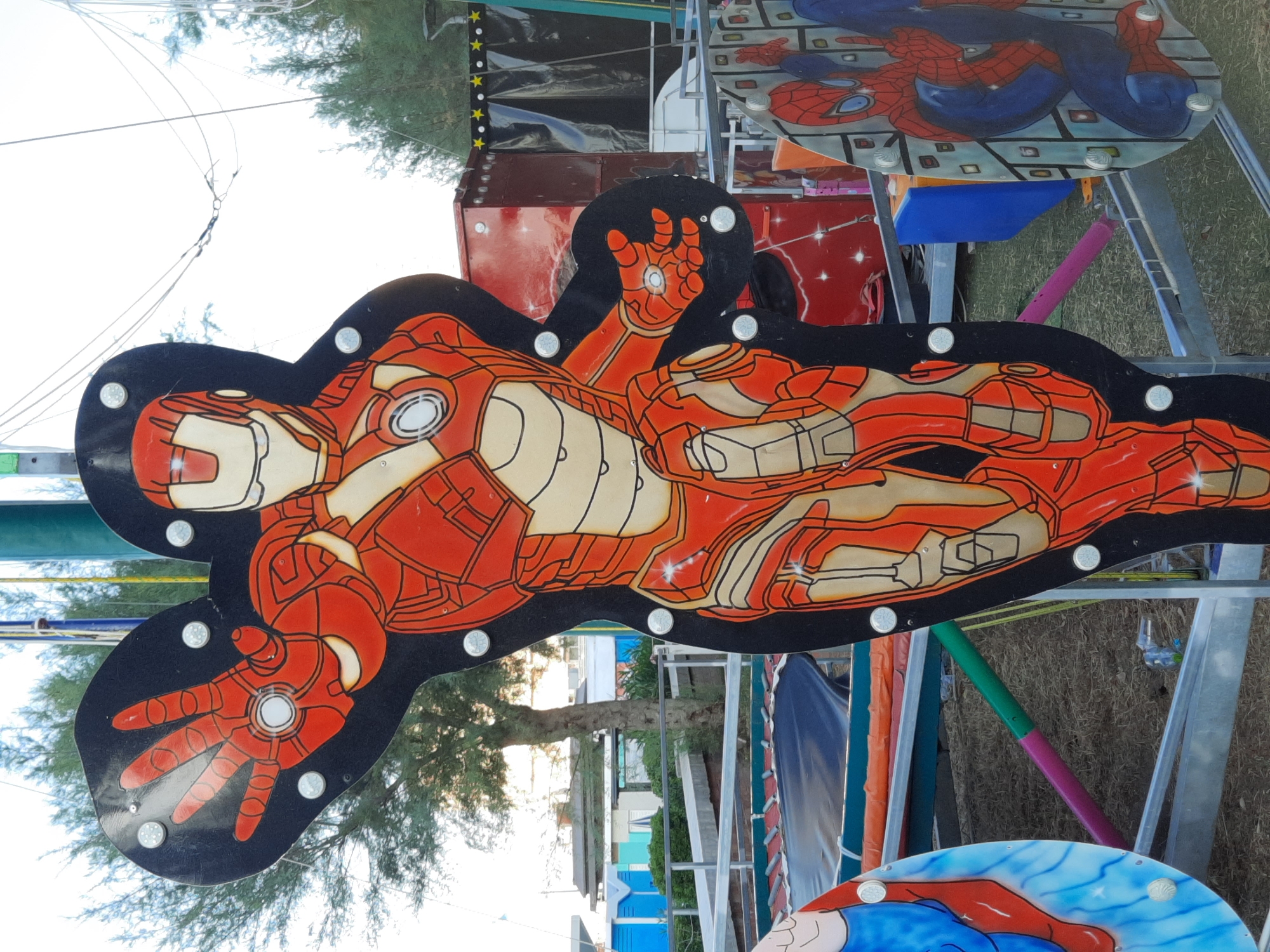 This is a full, slightly low-angle, outdoor shot of a cut-out figure of Iron Man, a superhero. The figure is vibrant orange and cream, with a black background. It is attached to a metal structure that appears to be part of a fairground ride or attraction. Several silver circular fasteners are visible, holding the figure to its backing. Iron Man's left hand is raised, palm facing the viewer, and his right hand is slightly lowered, also facing outwards. The figure's armor is detailed with lines and shading.
To the left of the Iron Man cut-out, a portion of another cut-out featuring a character, likely Superman given the blue and red colors, is visible. This figure is also attached to the same metal structure.
In the background, several elements suggest an outdoor amusement setting. A blue and orange trampoline-like structure is visible beneath the cut-outs. Further back, a red rectangular structure with a starry pattern is situated behind the Iron Man figure. A dark tarp or curtain hangs behind the red structure, and beyond that, trees and what appear to be beach huts or small buildings are discernible. The sky is bright and overcast.
The overall scene suggests a temporary setup in a public recreational area, possibly a seaside promenade or a fairground. The presence of beach huts and trees points towards a coastal location. Based on the visual cues, this scene is likely located in Pesaro, Italy, a coastal city known for its beaches and amusement areas.