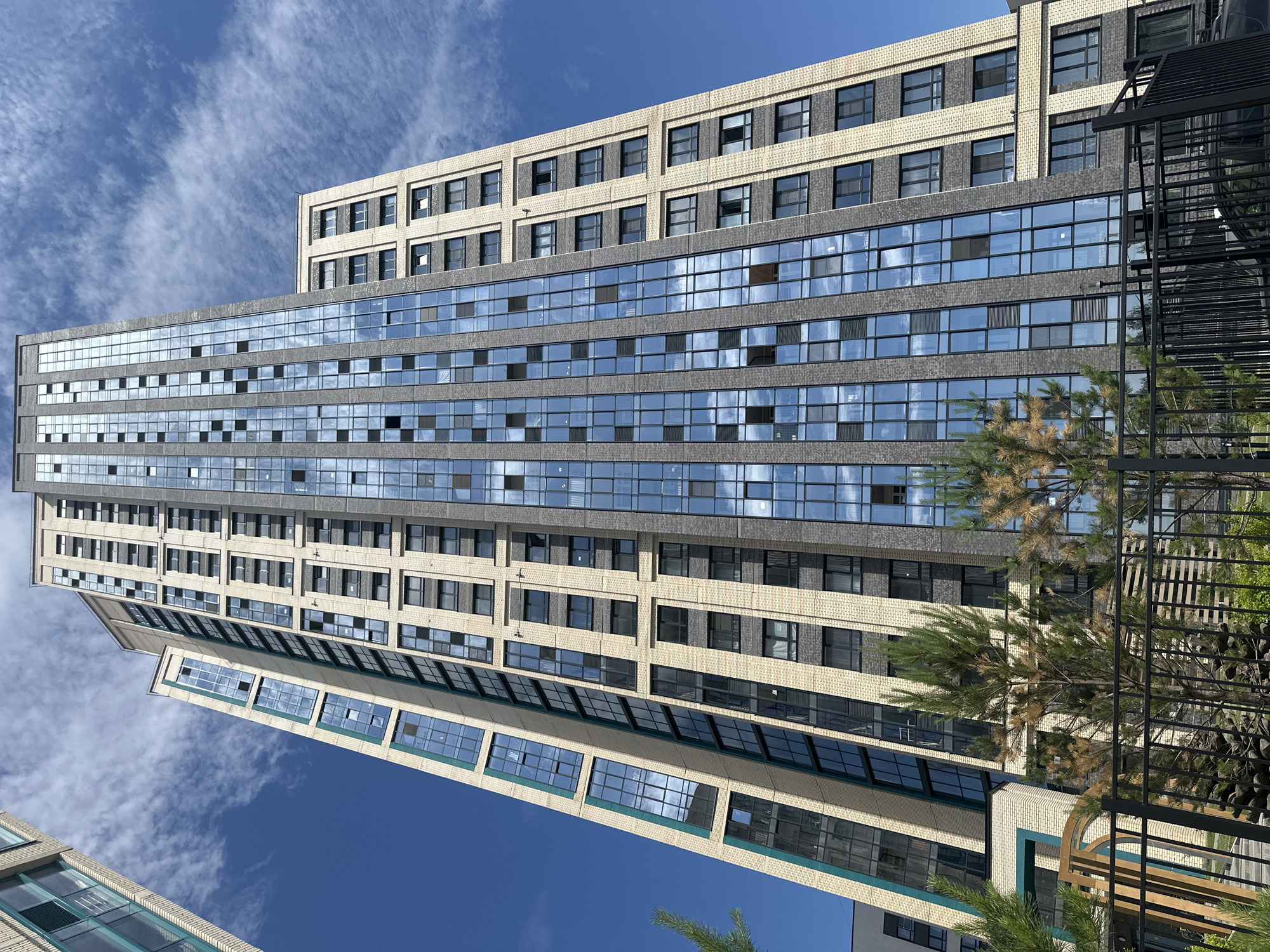 A low-angle, full shot captures a modern, tall apartment building with a blue sky and white clouds. The building is composed of two main sections: a section with a light beige brick facade and dark gray window frames, and a section with dark gray stone cladding and large blue-tinted glass windows. The glass windows reflect the sky and clouds. The building has a stepped design with protruding sections. In the foreground, a black metal fence runs horizontally across the bottom of the image, with some greenery and trees behind it. The trees are sparse and appear to be pine trees. A partial view of another modern building is visible on the left side of the image.