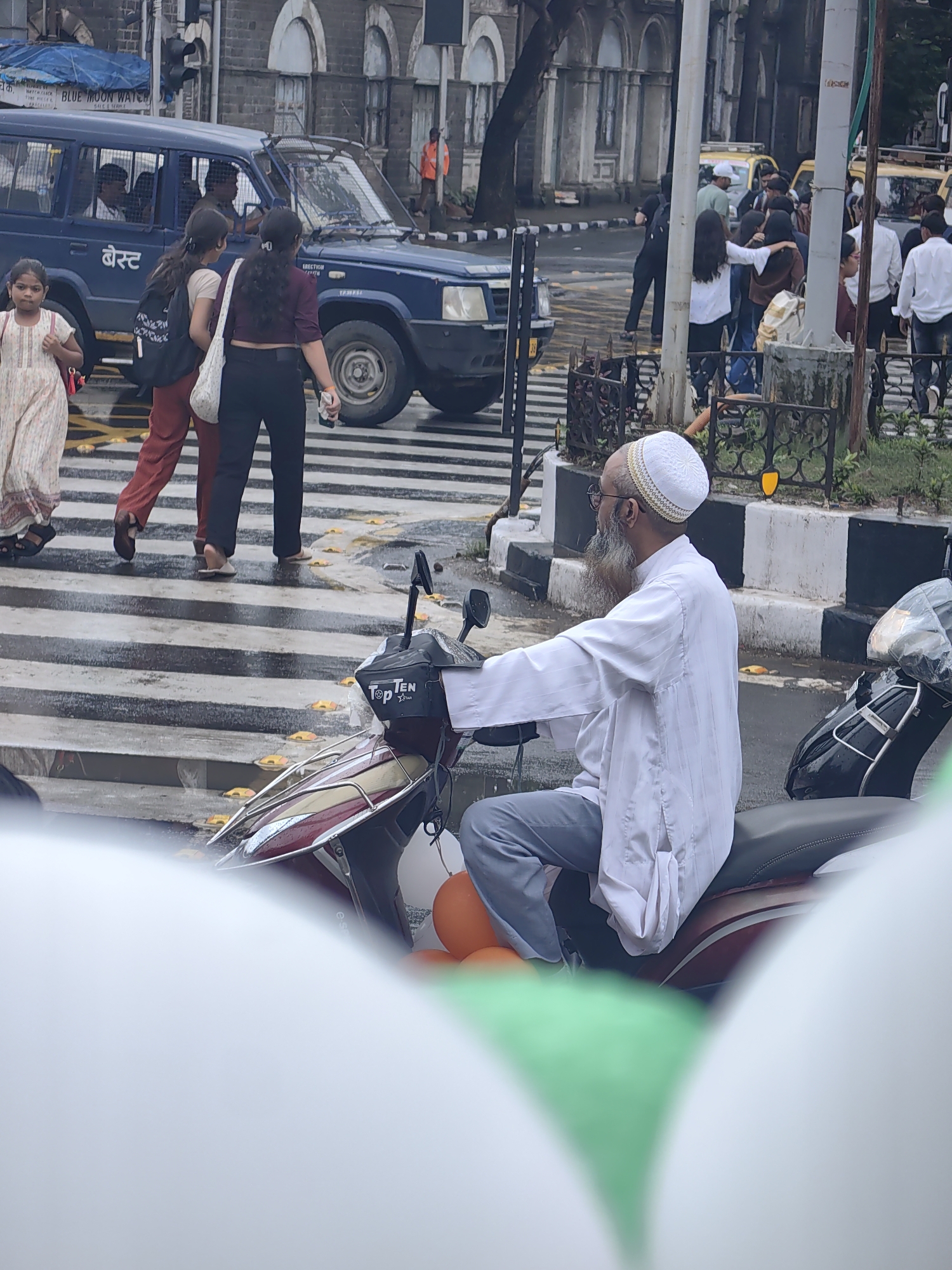 An overhead shot captures a busy street scene in Mumbai, India. In the foreground, a man with a white cap and beard rides a scooter, his body angled towards the right of the frame. He wears a white kurta and gray trousers. In front of him, balloons in orange, white, and green add a festive touch.
Further back, a group of people are crossing a street at a zebra crossing. Three women, two in maroon pants and a white top, and one in black pants and a maroon top, walk together. A young girl in a cream-colored dress stands to the left, looking towards the side. Behind them, a dark blue police vehicle is parked, its windows tinted.
The street appears wet, suggesting recent rain. Traffic signs and a yellow taxi are visible in the background, hinting at the urban environment of Mumbai. The architecture in the background is classic, with arched windows and pillars, indicating a historic part of the city.