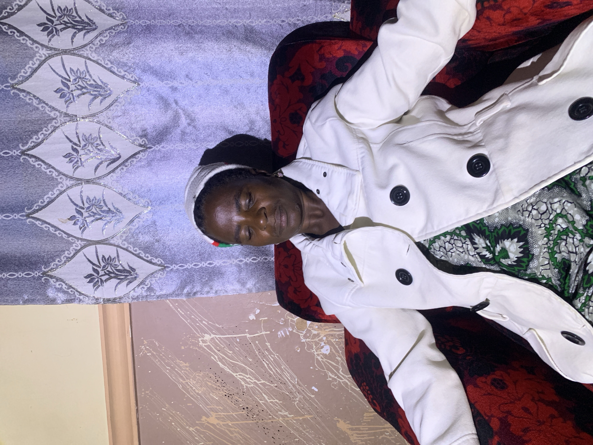 A Black woman wearing a white jacket and a head covering is seated in a red upholstered chair. The chair is positioned in front of a grey curtain with a floral pattern. To the left of the curtain, a wall painted in a light color is visible, with splotches of white and beige paint splattered across it. The woman's eyes are closed, and she is looking downwards. The scene appears to be an indoor setting in Barakin Sabawa, Nigeria.