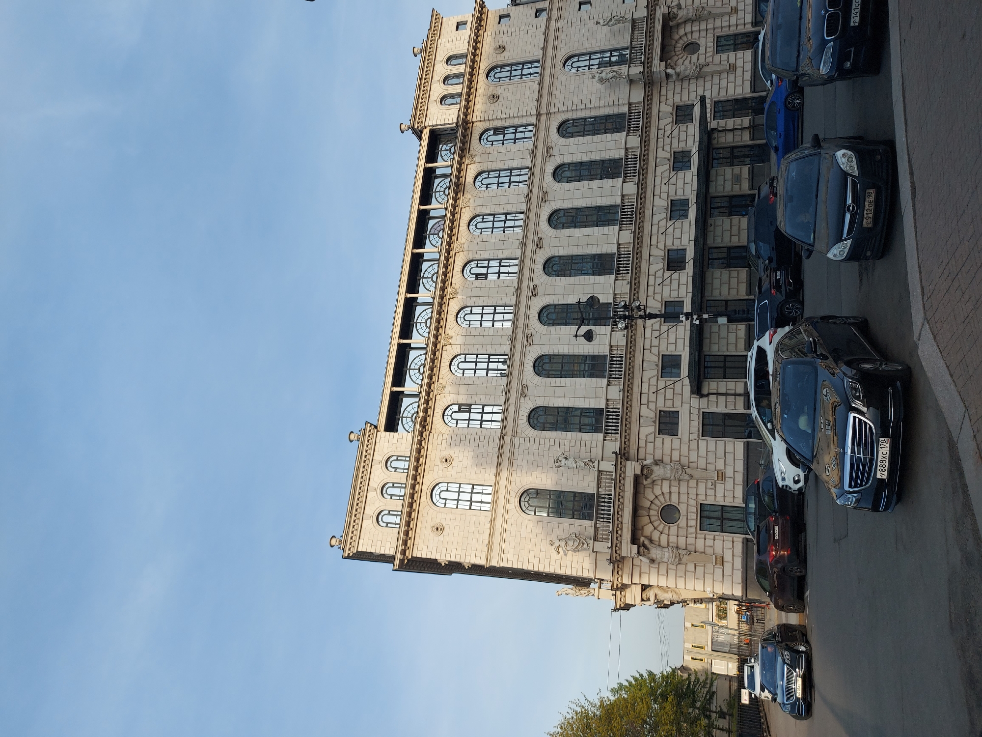 The image depicts a large, ornate building in Saint Petersburg, Russia. The building is characterized by its light-colored facade, multiple stories, and rows of arched windows. Architectural details include statues and decorative elements. Parked cars line the street in front of the building, including a Mercedes-Benz with the license plate "Y888XC 178". The sky is clear and blue. The scene appears to be daytime, likely early morning or late afternoon due to the angle of the light.
