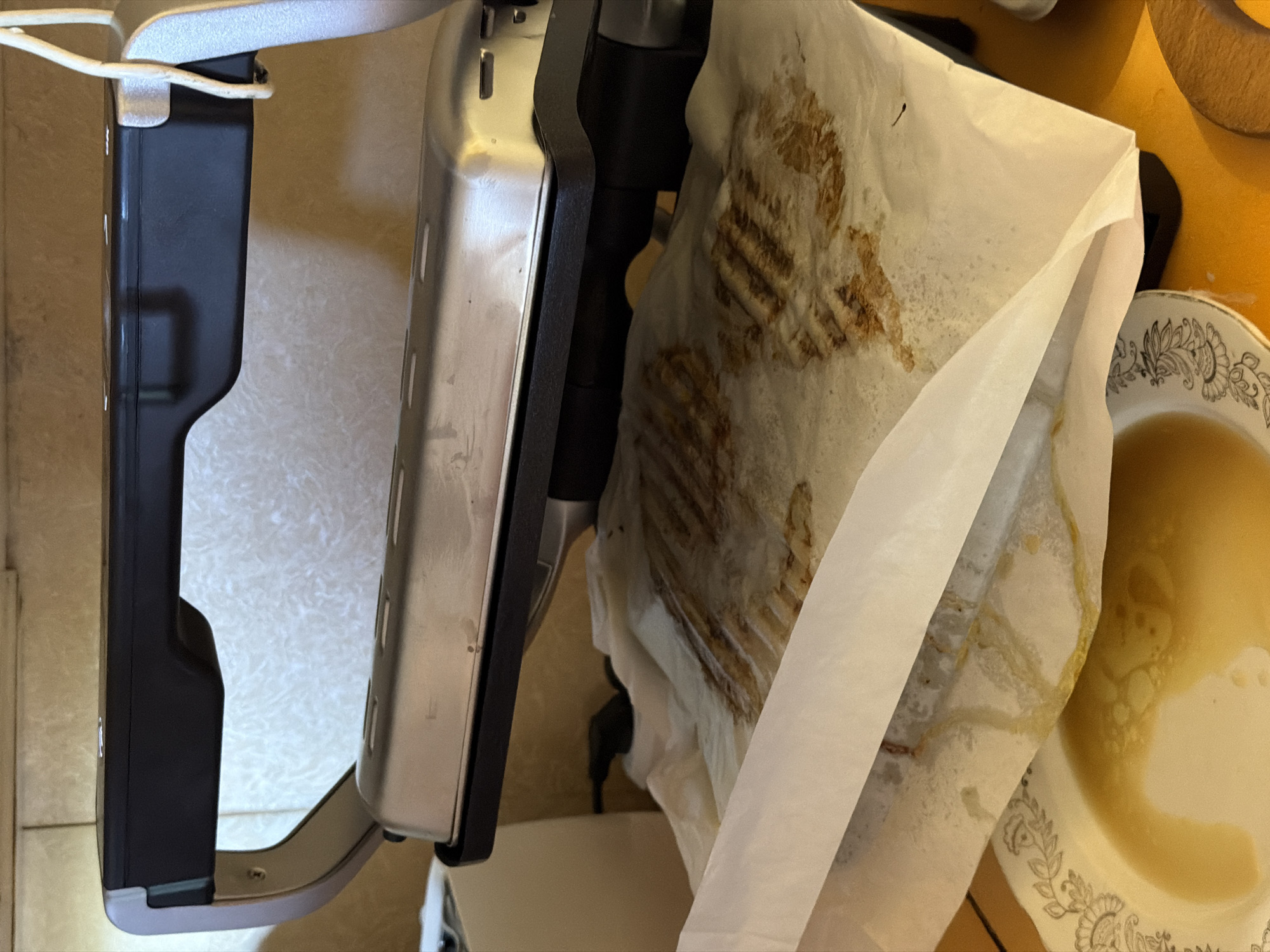 The image shows a close-up, top-down view of a waffle maker with partially cooked waffles. The waffle maker is open, revealing the top and bottom grates. The waffles are on parchment paper, which is laid out on a yellow surface. The waffles are a golden brown color and have a distinct waffle pattern. Some of the waffles appear to be sticking to the parchment paper. To the right of the waffle maker, there is a plate with some liquid, possibly oil or syrup, in it. The plate has a decorative floral pattern. The background is a neutral color, possibly a countertop. The lighting suggests it is daytime. There is no visible text in the image. The setting appears to be a kitchen. The image is a bit blurry, suggesting it may have been taken with a mobile phone. The activity taking place is the cooking of waffles.