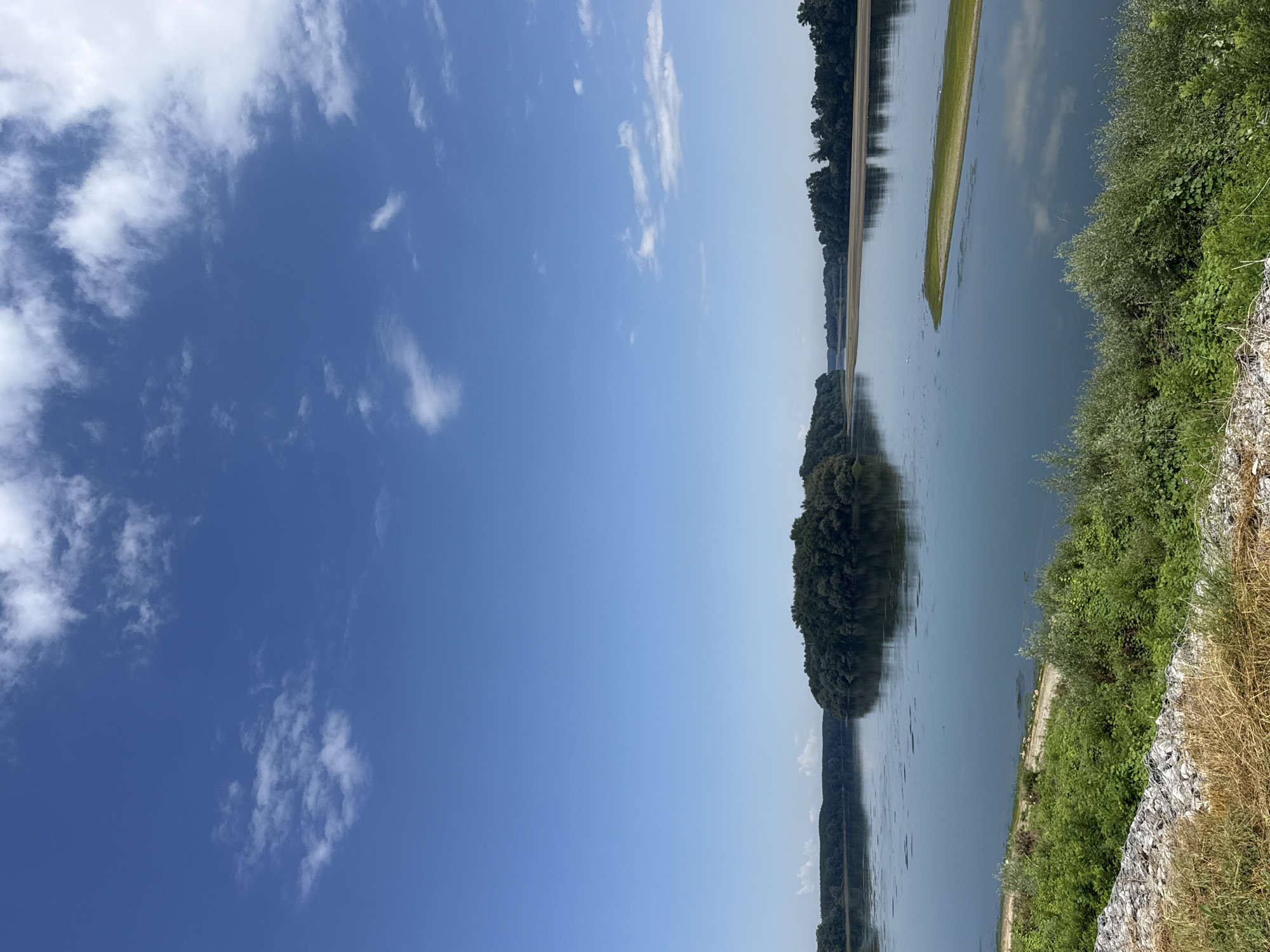 The image depicts a wide, calm river with a distinct island covered in dense green trees in the center. The sky is a clear, bright blue with scattered white clouds. The riverbanks are lined with lush green vegetation, and in the foreground, there's a rocky embankment with dry grass. The overall scene suggests a serene natural landscape. This view is consistent with the landscapes along the Danube River near Belene, Bulgaria, known for its islands and riparian ecosystems.
