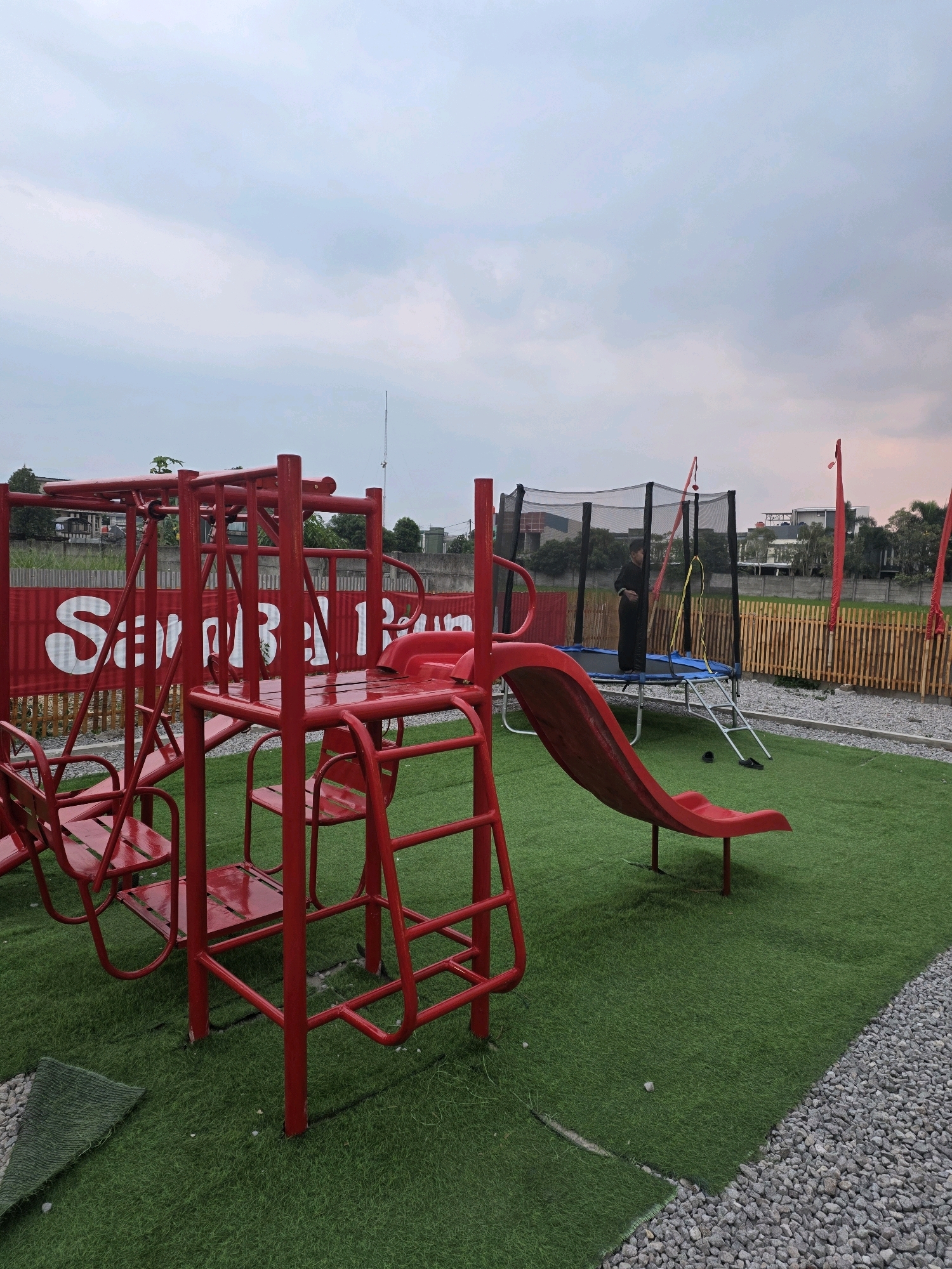 A red metal playground structure is in the foreground of this outdoor scene, featuring swings and steps. To the right of the playground, a red slide descends onto green artificial turf. Further back, a trampoline with a black net is visible, and a young boy in dark clothing stands on it. A wooden fence borders the play area, with buildings and trees visible beyond it. A red banner with white text is stretched across the background. The ground is covered in a combination of artificial turf and gravel. The sky is overcast with hints of pink and orange, suggesting it is either dawn or dusk. The image appears to be taken in the city of Bandung, Indonesia, given the visual context of the surrounding landscape and architecture.