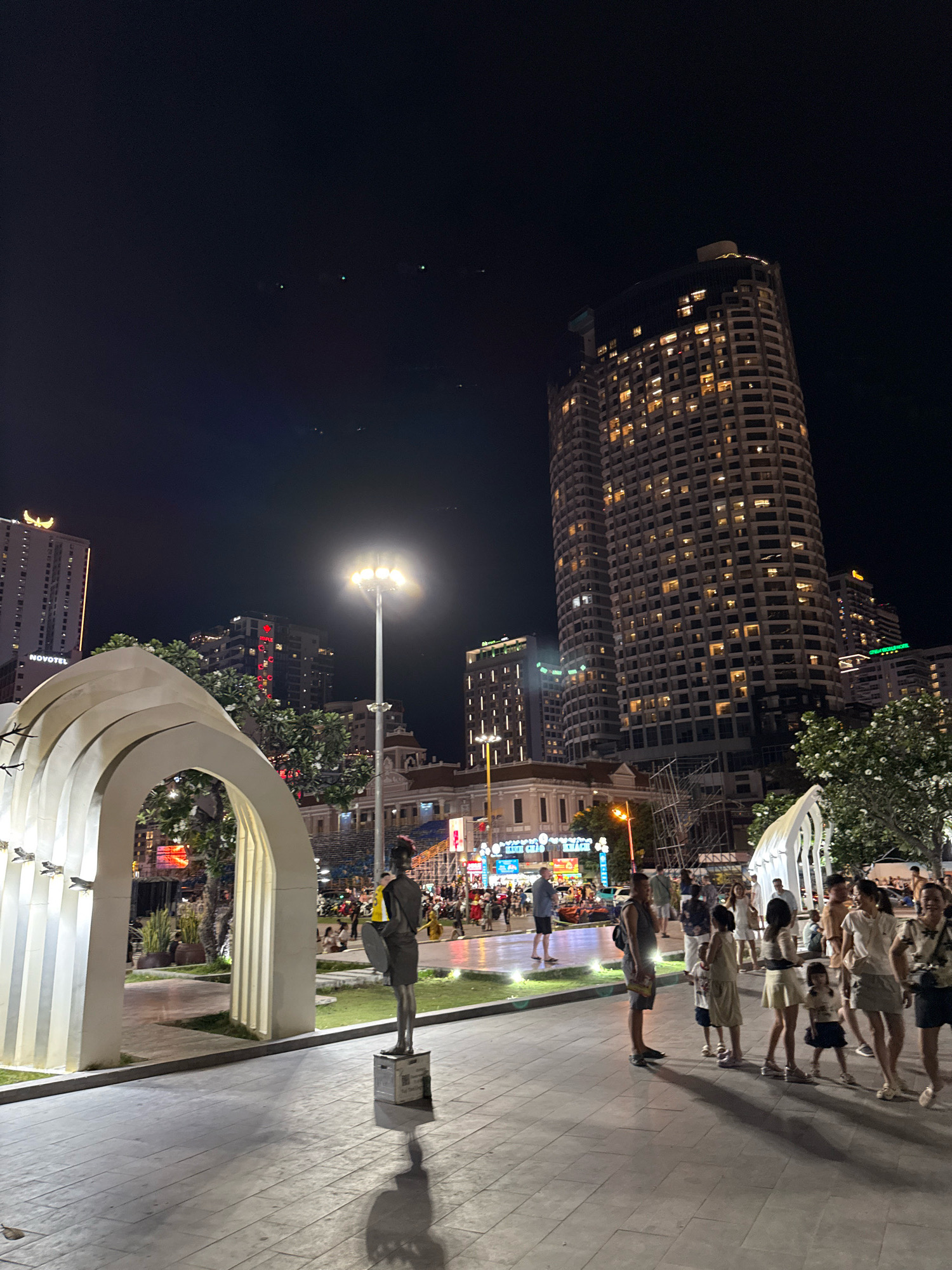 The image captures a nighttime scene in Nha Trang, Vietnam, likely along Tran Phu Street, a popular seaside promenade. In the foreground, a contemporary white archway structure stands on a paved walkway. To the right of the archway, a dark, full-body statue of a person holding a circular object stands on a square pedestal. Beyond the archway and statue, a grassy area is illuminated by soft lights, and a street lamp with multiple bulbs illuminates the scene further.
In the background, several multi-story buildings are visible. The most prominent is a tall, cylindrical building with numerous windows, many of which are lit, suggesting it's a hotel or residential tower. Other buildings, varying in height and architectural style, line the street, some displaying illuminated signage. The sky above is dark, with a few distant lights possibly representing aircraft or stars.
A diverse group of people, including adults and children, are present in the middle ground and background, strolling, standing, and interacting. Their attire suggests a warm climate. The overall atmosphere appears to be one of public recreation and urban nighttime activity.