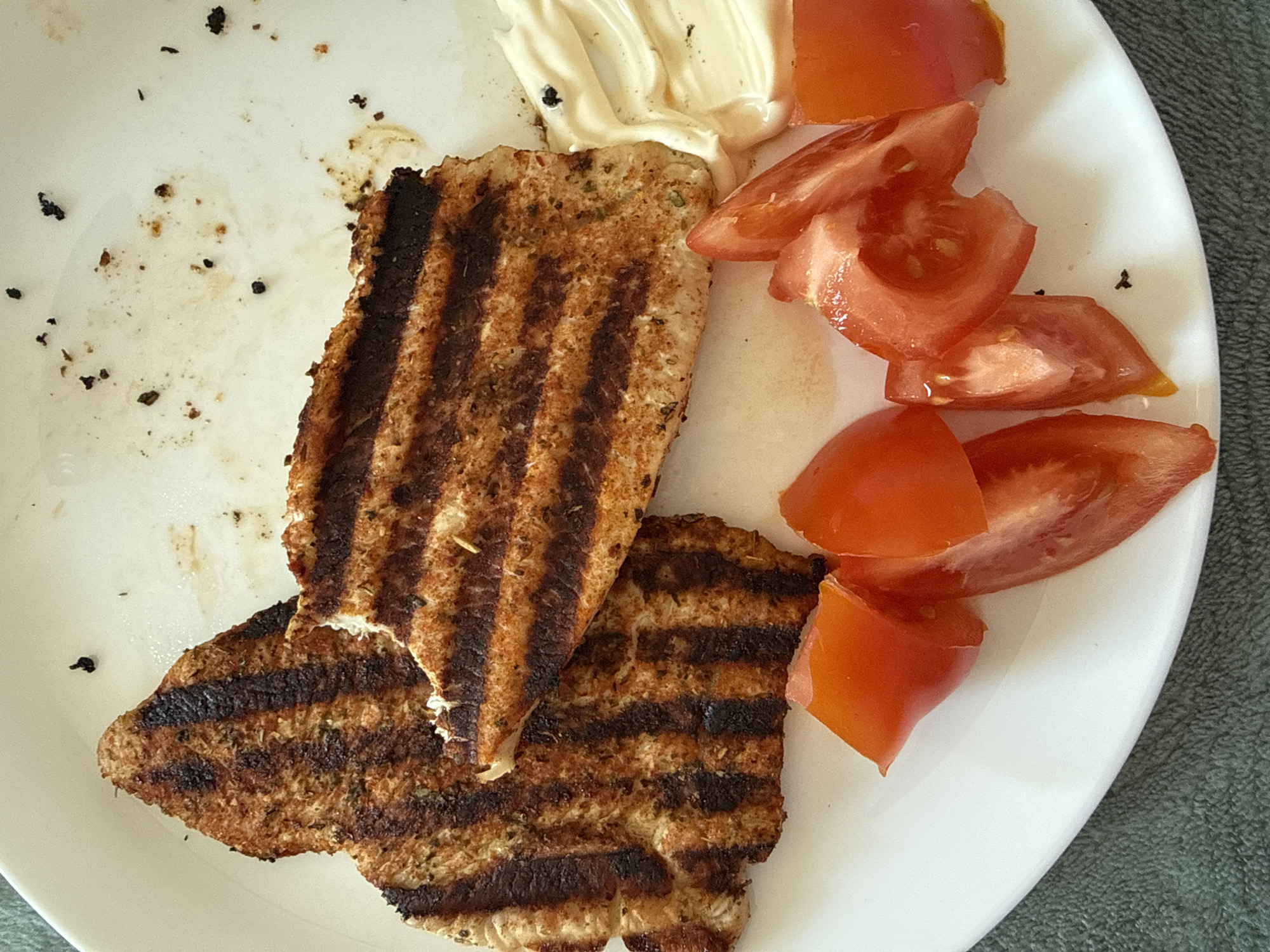 A white plate contains two grilled slices of bread with grill marks, a dollop of white sauce (possibly mayonnaise), and several slices of tomato. There are some crumbs and dark specks scattered on the plate. No people are visible. The scene appears to be a simple meal or snack. There is no indication of Izhevsk, Russia or any city, in the image.