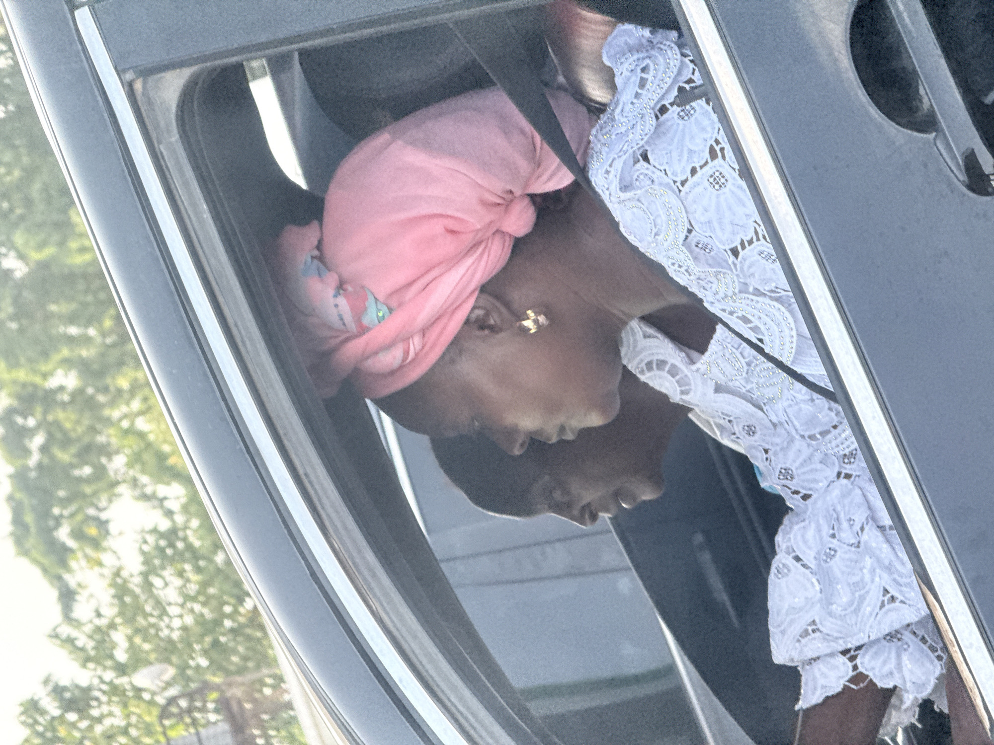 An outdoor, brightly lit image captures a close-up view of a person leaning out of a dark-colored car window, likely in Iddo Tudunwada, Nigeria. The primary subject is a dark-skinned woman seen in profile, wearing a vibrant pink headwrap, possibly a gele or turban. She has a distinctive gold earring in the shape of a cross or a similar symbol. Her attire includes an ornate white top or dress, richly textured with a lace or embroidered pattern featuring shiny accents.
The woman is looking intently towards the left of the frame, outside the car. A clear reflection of another dark-skinned individual, also in profile, is visible on the lower part of the car's window or body panel, positioned below and slightly behind the main subject, suggesting another passenger or a distinct reflection of the woman herself.
Through the car window and beyond its frame, blurred green foliage from trees or bushes indicates an outdoor, natural setting. The bright, even lighting suggests it is daytime, possibly morning or afternoon, with clear weather. The image contains no visible text.