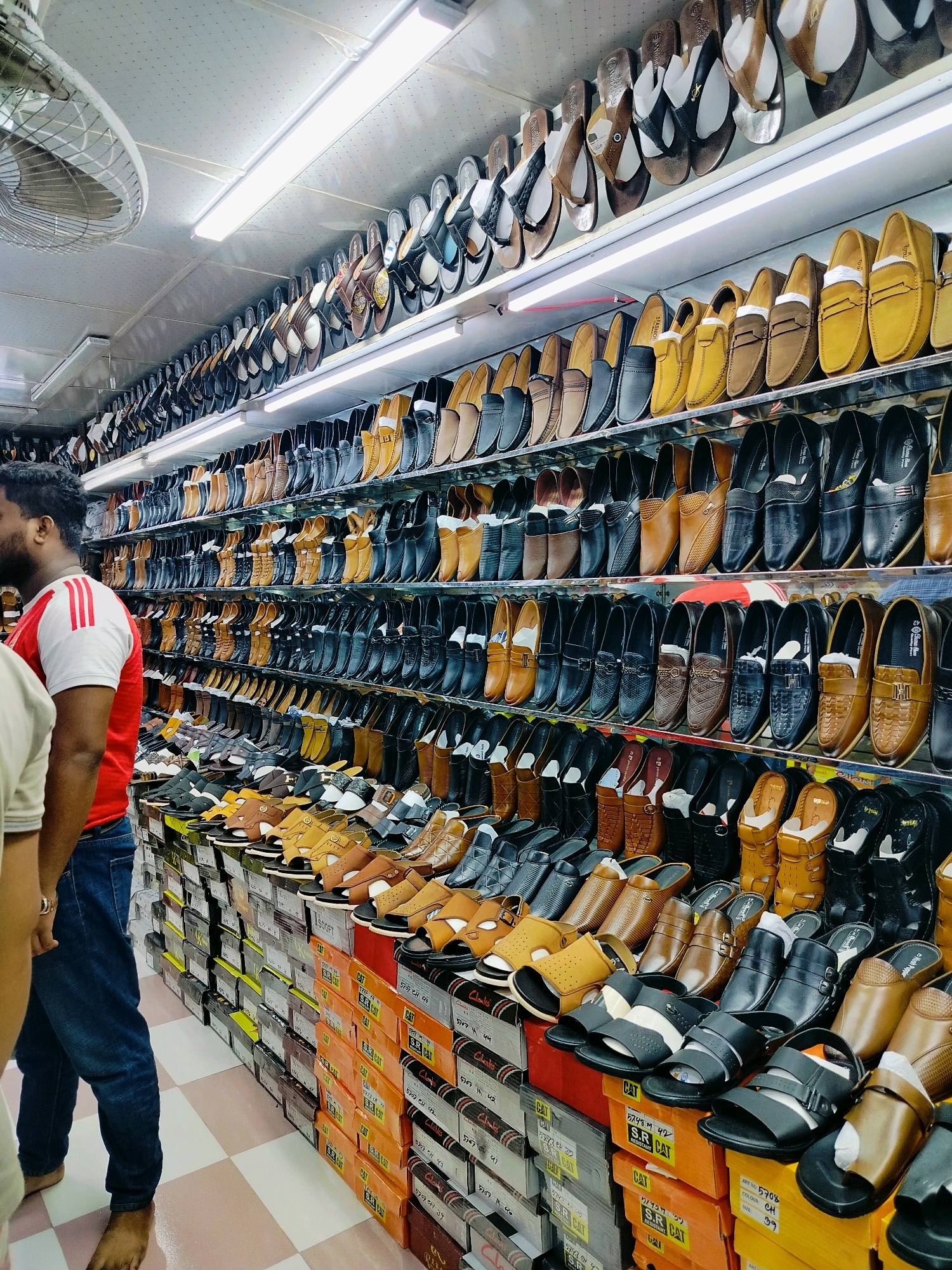 In a retail store in Dhaka, Bangladesh, a wide assortment of men's footwear is displayed on multiple shelves and stacked in boxes. The footwear includes a variety of styles such as loafers, sandals, and slip-on shoes, presented in colors like black, brown, tan, and yellow. The store appears well-lit with fluorescent lighting, and a ceiling fan is visible in the upper left corner. A man wearing a red and white striped shirt and blue jeans is standing on the left side of the frame, facing away from the camera, and another person is partially visible to his left. The floor is tiled with a checkered pattern of white and pink squares. The sheer volume of shoes suggests a commercial establishment catering to a large customer base.