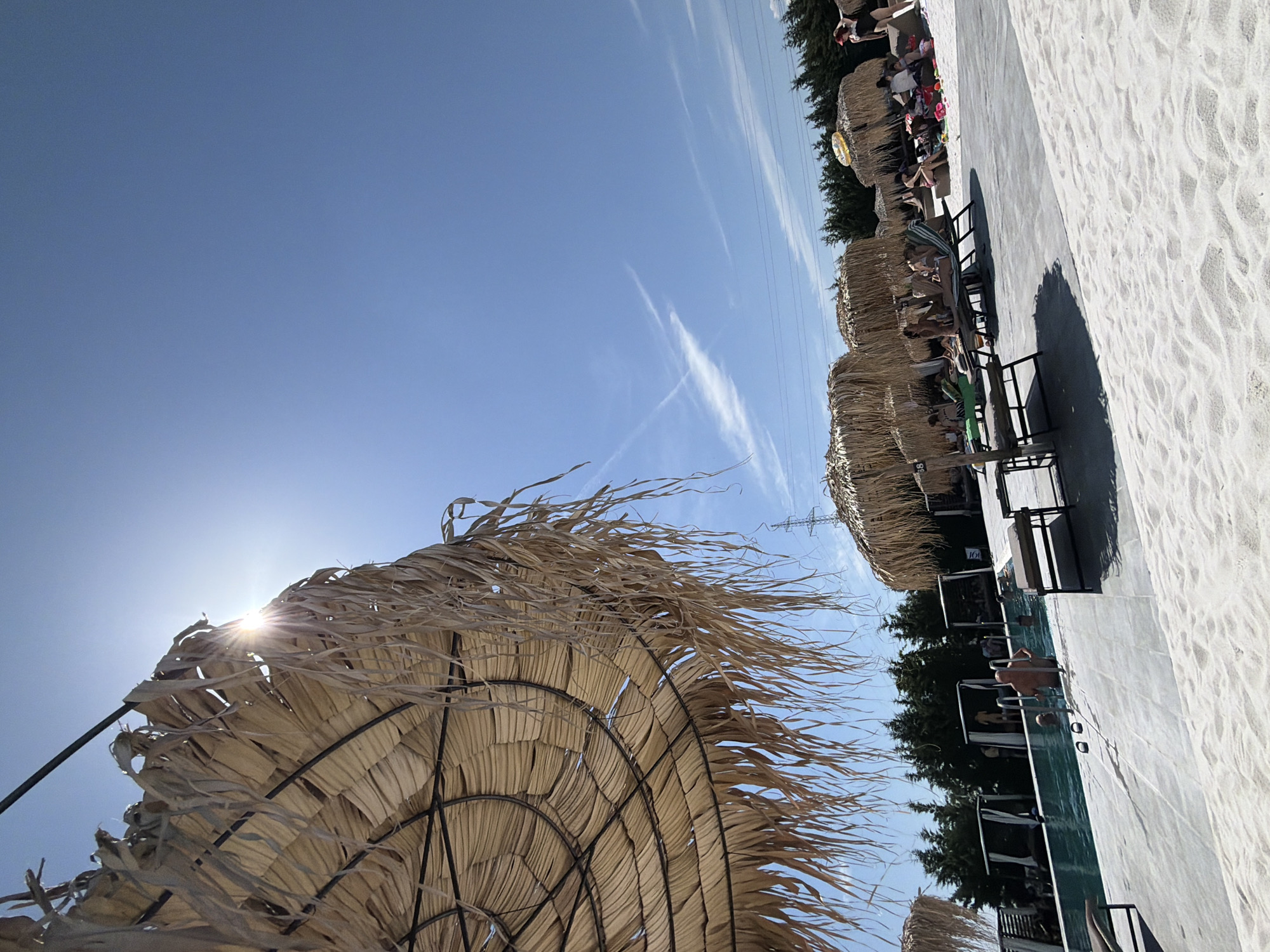 The image depicts a sunny day at a resort in Kazichene, Bulgaria. In the foreground, a large straw umbrella provides shade. Beyond it, a swimming pool is visible with several people in it. Sun loungers and more straw umbrellas are arranged on a sandy area next to the pool. In the background, trees form a dense line, and power lines are strung across the sky. Several individuals are seen relaxing on loungers and near the pool. The overall scene suggests a leisurely atmosphere at a beach resort.