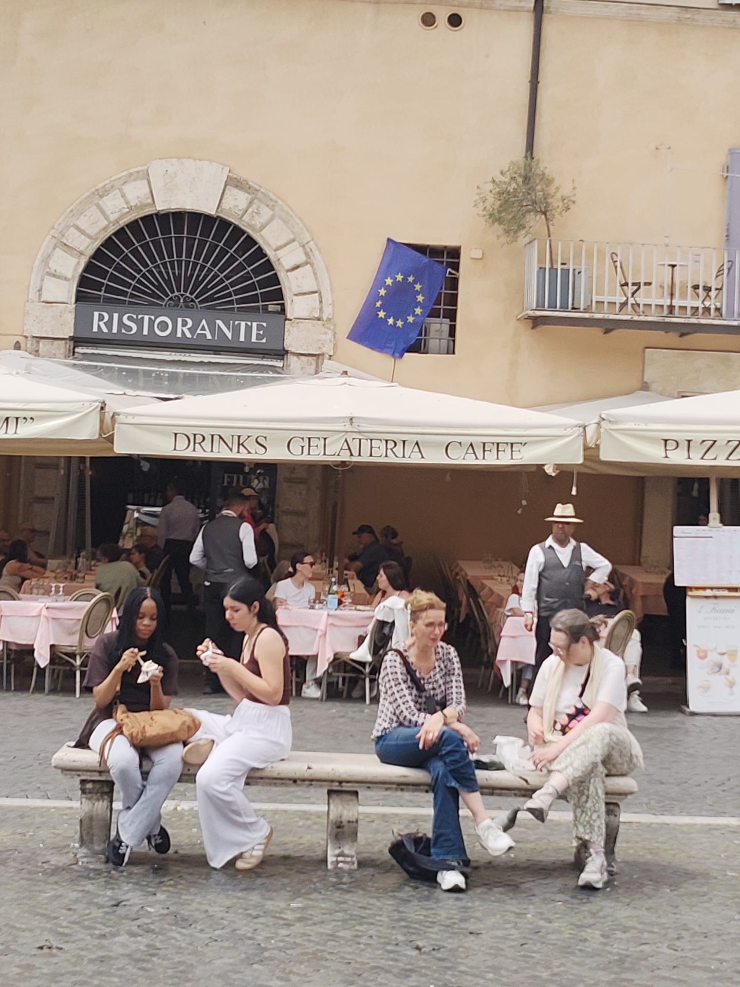 The image shows a street scene in Rome, Italy. In the background is a building with a restaurant called "Ristorante". The restaurant has a striped awning labeled "DRINKS GELATERIA CAFFE' PIZZA". Diners are seated at tables under the awning, being served by waiters. A man in a straw hat and vest stands observing the scene. In the foreground, four women are seated on a stone bench. Two women on the left appear to be eating gelato. The woman next to them has on a patterned blouse and jeans. The fourth woman wears patterned pants, a white shirt, and a scarf. A European Union flag is visible on the side of the building.