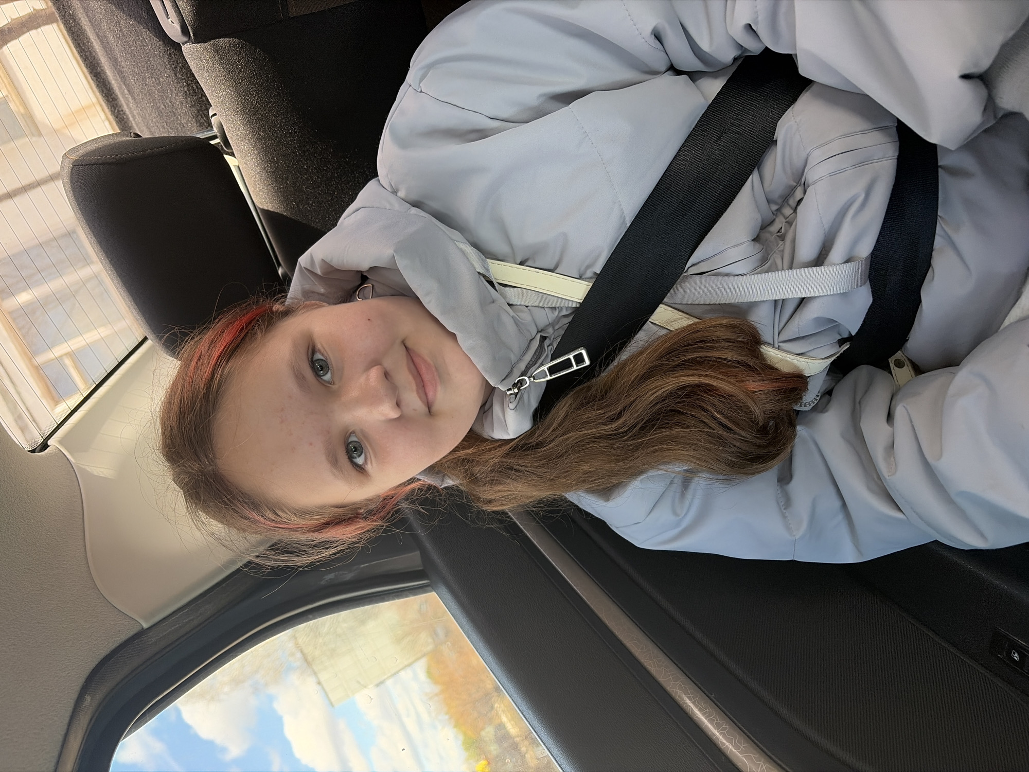 A young person with light skin, light eyes, and brown hair with red highlights is sitting in the back seat of a car. They are wearing a light gray puffy jacket and are secured with a black seatbelt. A light-colored strap hangs from the jacket. The interior of the car is dark, and outside the window, one can see a blurry view of trees and a blue sky with clouds. The location is Unknown, Unknown.