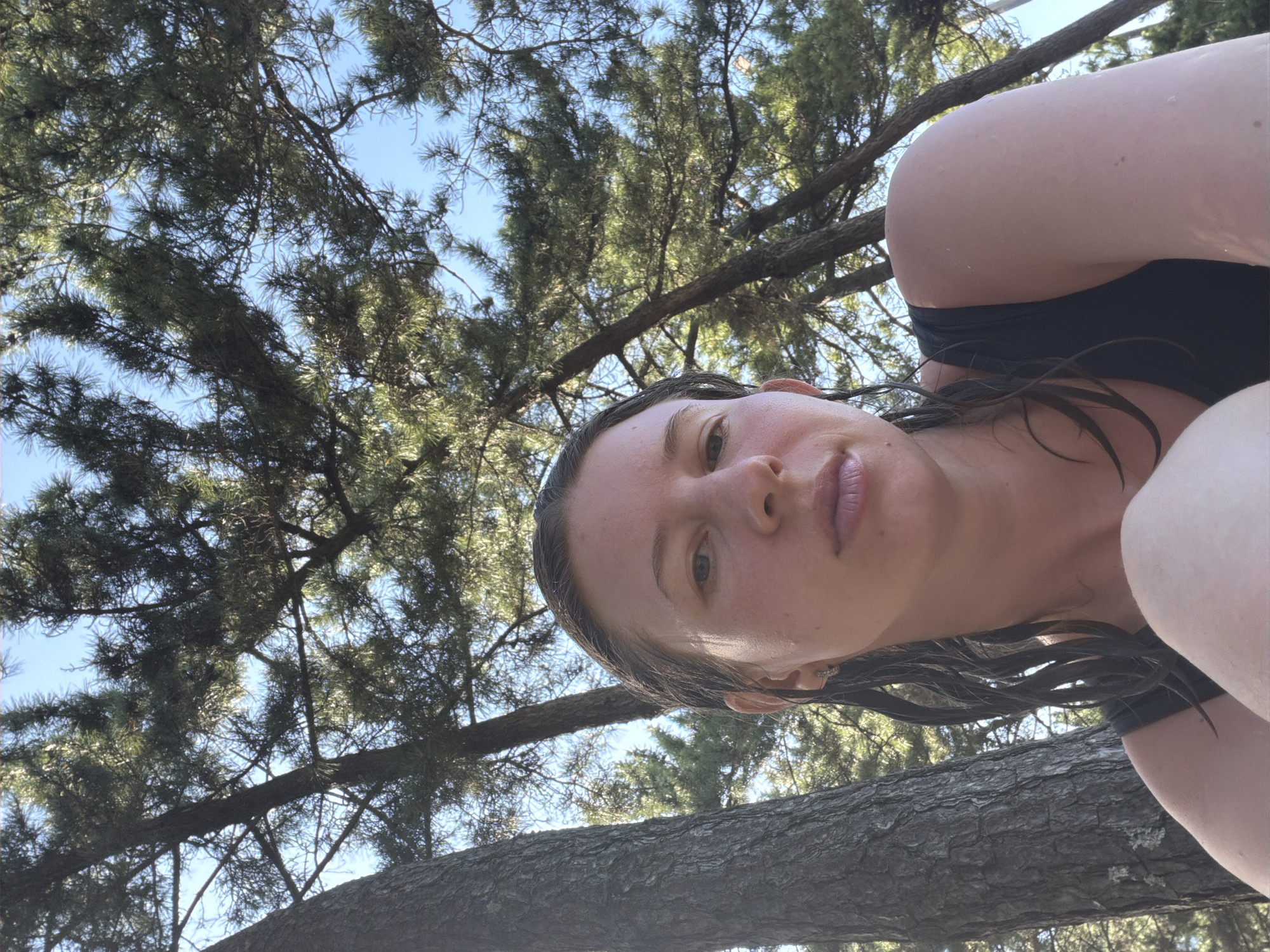 A woman with wet hair, wearing a black tank top, poses for a selfie outdoors. She is seated in front of trees and a clear blue sky, suggesting a natural setting like a park or forest. The woman's face is the primary focus, with her hair framing her features. The background consists of pine trees, their branches reaching towards the sky, indicating a possible location in a coastal area like Gangneung-si, South Korea, known for its pine forests and beaches. The lighting suggests it is daytime, with sunlight filtering through the leaves.