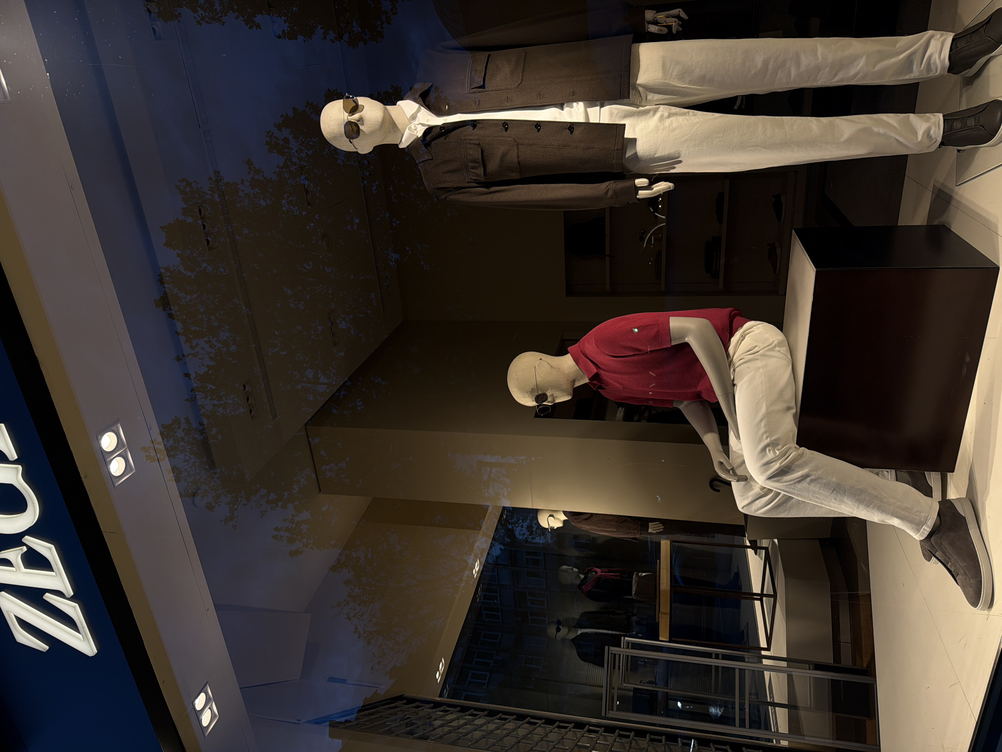 The image depicts a ZARA store window display in Sofia, Bulgaria. Two mannequins are positioned at the top, one standing and wearing a brown jacket over a white shirt and light-colored pants, and the other prone. Below them, another mannequin is seated and wearing a red polo shirt and light-colored pants. The store window reflects the cityscape and trees outside, along with the store's branding.