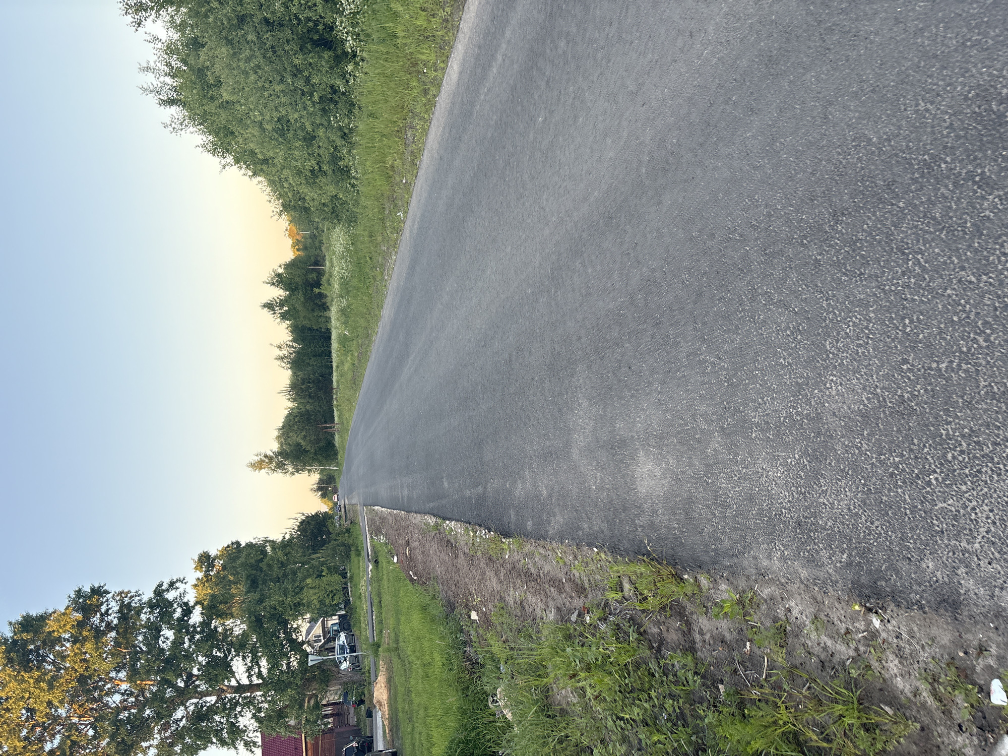 The media file presents a long, straight stretch of newly paved asphalt road extending into the distance under a vast sky. The scene is set in what appears to be a rural or semi-rural area, characterized by abundant greenery.
On both sides, the road is flanked by unkempt grass and dirt shoulders, with some loose debris visible along the edges, suggesting recent construction or grading work. To the left side of the road, a dense line of lush green trees forms a natural boundary. Further beyond these trees, in the mid-ground, a residential area can be discerned, featuring several houses with visible roofs and what appear to be parked vehicles, possibly cars or trucks.
The right side of the road also features greenery, with a line of trees extending further into the background, though less dense than those on the left. The sky above transitions smoothly from a light blue directly overhead to a warm, soft yellow or orange hue near the horizon, indicative of either early morning (sunrise) or late afternoon/early evening (sunset). The weather appears clear and calm, with no clouds visible.
There are no people or animals visible in the image. The overall impression is one of a quiet landscape, highlighting new infrastructure against a natural backdrop. No visible text is present in the image.