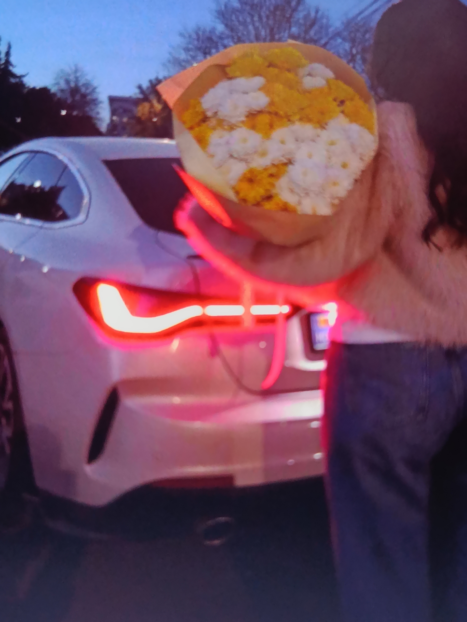 A person stands next to the rear of a white car in Bălți, Moldova. The car's taillights are illuminated with a bright red light. The person is holding a bouquet of yellow and white flowers. The background shows trees and buildings against a twilight sky. The image is slightly blurry.