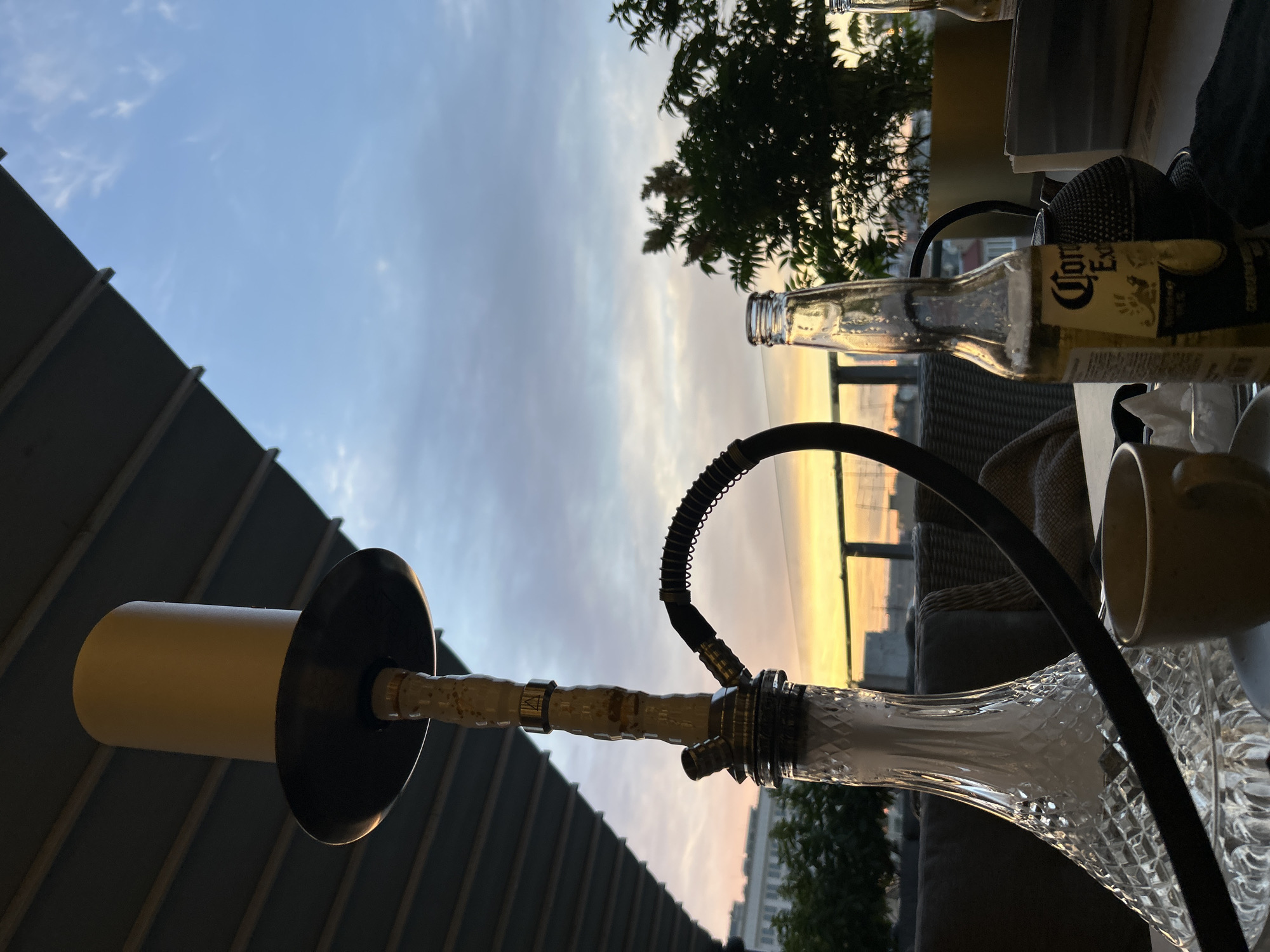 A clear glass hookah is the primary subject, positioned on a table outdoors, likely on a balcony or patio, with a dark-colored railing in the foreground. The hookah's stem is intricate, with a bamboo-like pattern and silver accents, leading to a black hose. A white ceramic bowl sits atop the stem. In the background, an empty Corona Extra beer bottle is visible, along with a white mug and part of a black mesh speaker.
The scene is set against a twilight sky, characterized by a gradient of soft blues and pale oranges, indicating either sunrise or sunset. Wispy clouds are scattered across the sky. Beyond the table, there's a hint of a cityscape with buildings visible in the distance. A lush green tree with small white flowers adds a natural element to the setting. The overall atmosphere appears relaxed and peaceful, perhaps a moment of quiet enjoyment after a day. The time of day is clearly dusk or dawn. There are no people visible in the image. The visible text on the bottle reads "Corona Extra". The location context of Novokuybyshevsk, Russia, is provided but not directly evident from visual cues within the image itself.