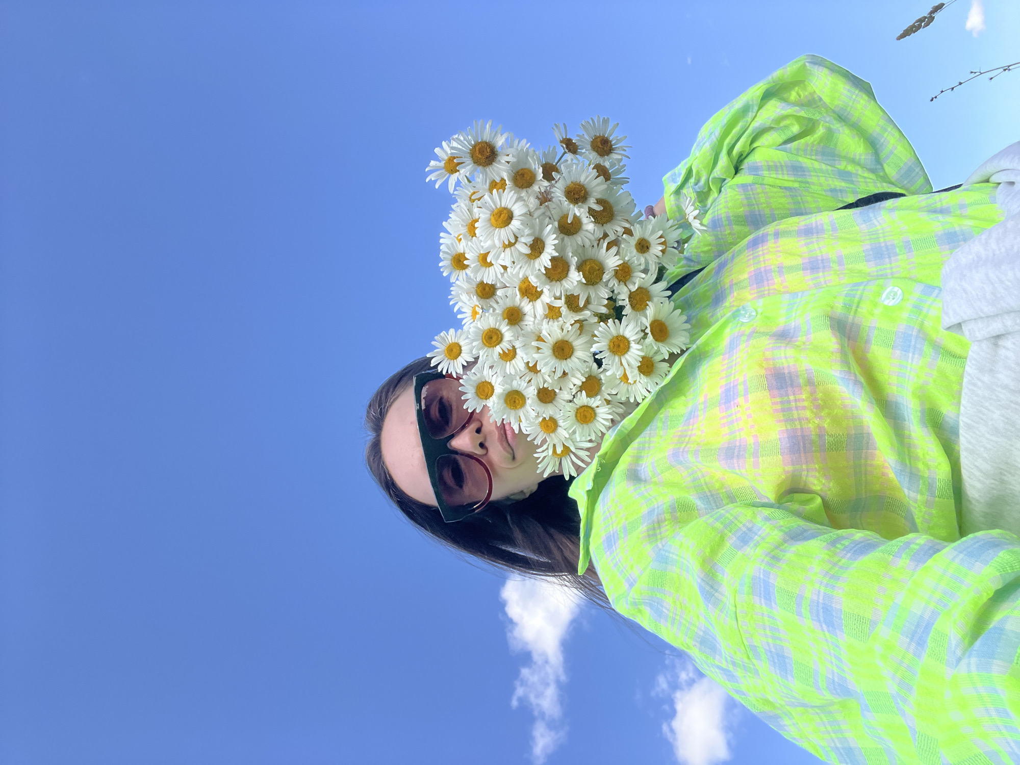 A low-angle, medium shot captures a person holding a bouquet of daisies against a bright blue sky with scattered white clouds. The individual, wearing dark-rimmed sunglasses and a neon green and blue plaid shirt, is positioned in the lower half of the frame. The flowers partially obscure their face. The background consists entirely of the sky and a hint of dry grass in the lower right corner. The location is identified as Pigozino, Russia.