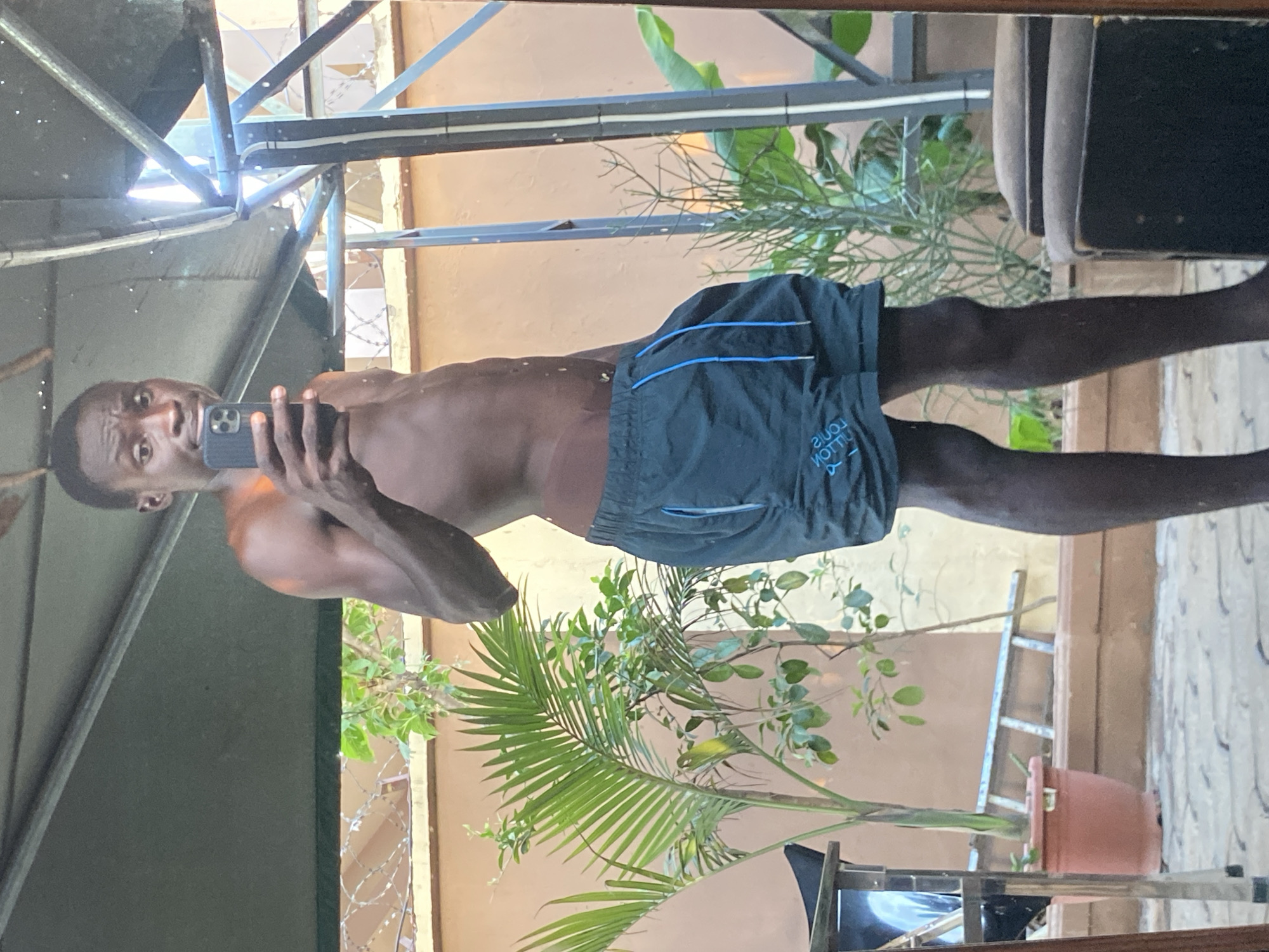 A shirtless man with dark skin holds a mobile phone and takes a selfie. He is wearing black shorts with blue stripes and a "Louis Vuitton" logo printed on them. He appears to be standing on a balcony or in a yard, possibly in Barden Yana, Nigeria. He is reflected in a mirror. Behind him, there is a view of plants in pots, a ladder, and a metal structure. Above, there is some metal framework and corrugated roofing. Barbed wire is visible on the left of the frame.