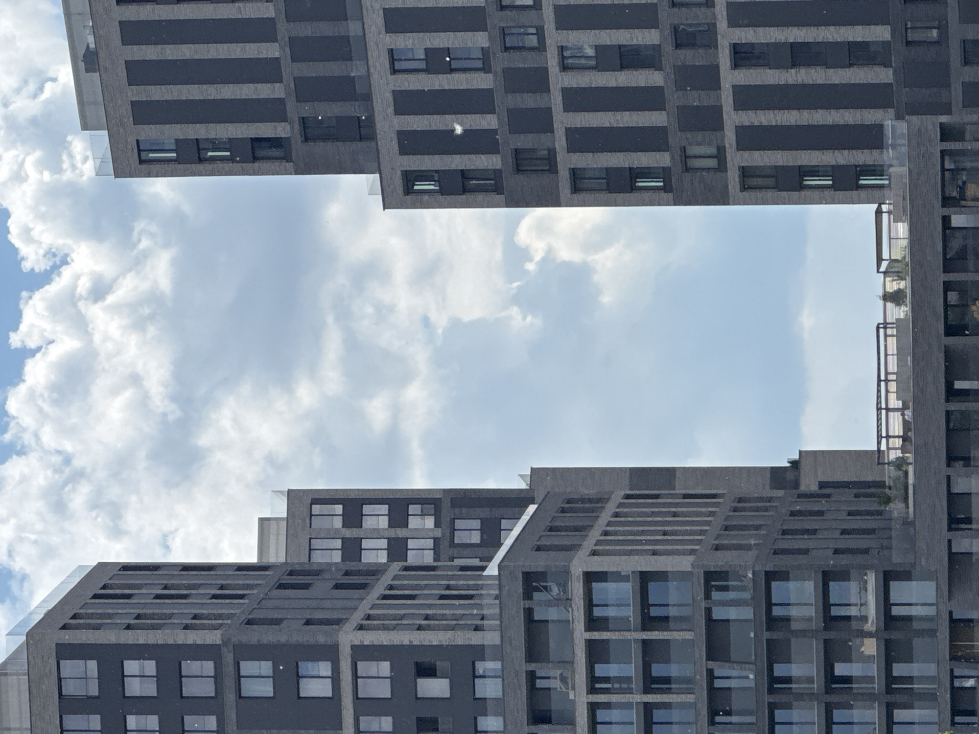 The image captures a modern architectural landscape against a bright, partly cloudy sky. Several contemporary buildings, predominantly dark gray or charcoal in color, are visible. The buildings exhibit a striking geometric design with numerous rectangular windows, many reflecting the blue sky and clouds.
In the upper part of the frame, a building features distinct horizontal bands that alternate with window sections, some appearing to be deeply recessed or cantilevered. A small, bright white object, possibly a feather or a piece of debris, is visible on one of these dark facade sections.
The lower portion of the image shows other buildings with a stepped or terraced structure, also dark-toned and adorned with multiple window arrays. Balconies with railings can be seen on the rightmost building, suggesting outdoor spaces. Transparent glass barriers are integrated into the design of some structures.
The sky, filling the central void between the buildings and serving as the background, is a clear blue punctuated by scattered, fluffy white cumulus clouds, indicating fair weather and bright daylight. No people or visible text are present in the image. The scene portrays a slice of modern urban environment, likely in Moscow, Russia.