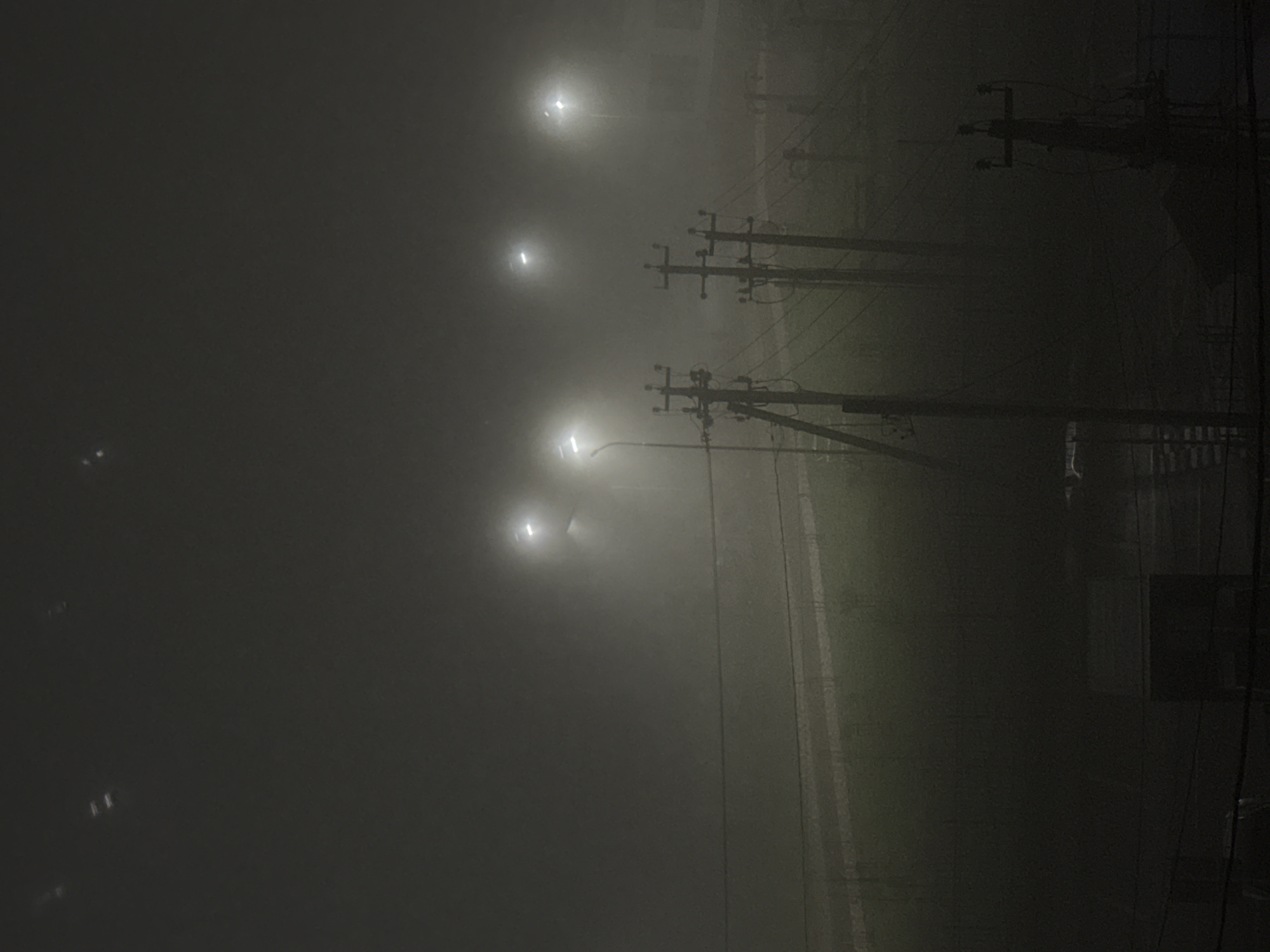 A dark, foggy cityscape at night. Several streetlights illuminate the scene with an intense glow. Rows of wooden power poles run along the roadside. Wires crisscross above, blurred by the heavy fog. A narrow strip of greenery is visible next to the road. A building is partially obscured in the upper background.