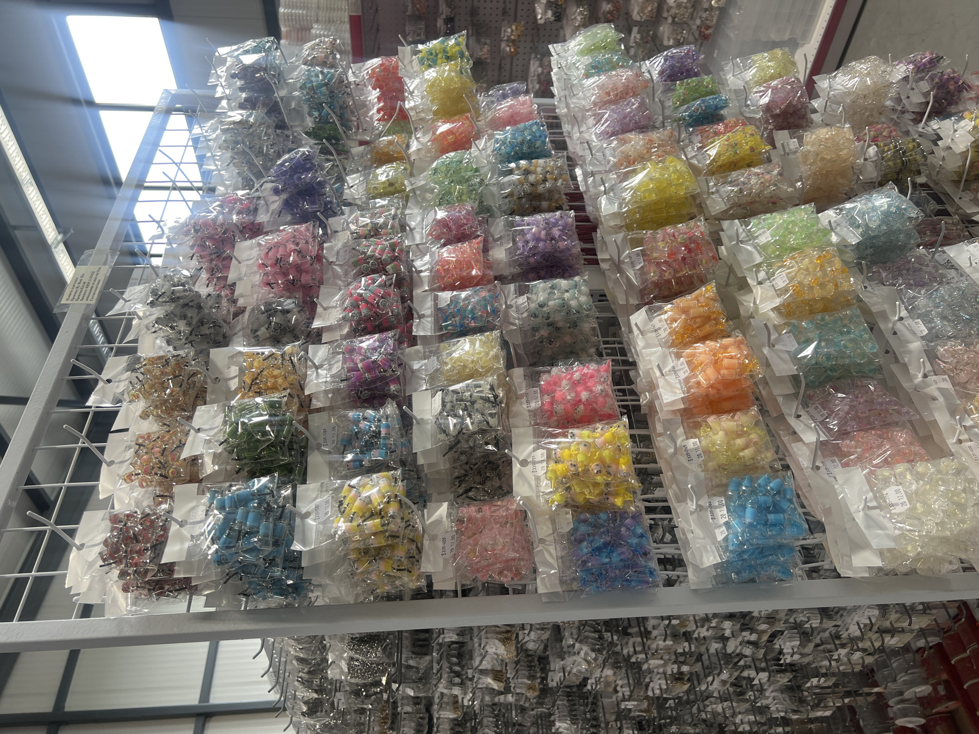 The image shows rows of individually packaged, colorful beads on a wire shelf. The beads are likely for sale. The packaging appears to be labeled with product codes and prices. The location is unknown, but based on the text in the OCR it is possibly the city of Burgas, Bulgaria. The environment is a retail or storage setting, indicated by the shelf and packaging style.
