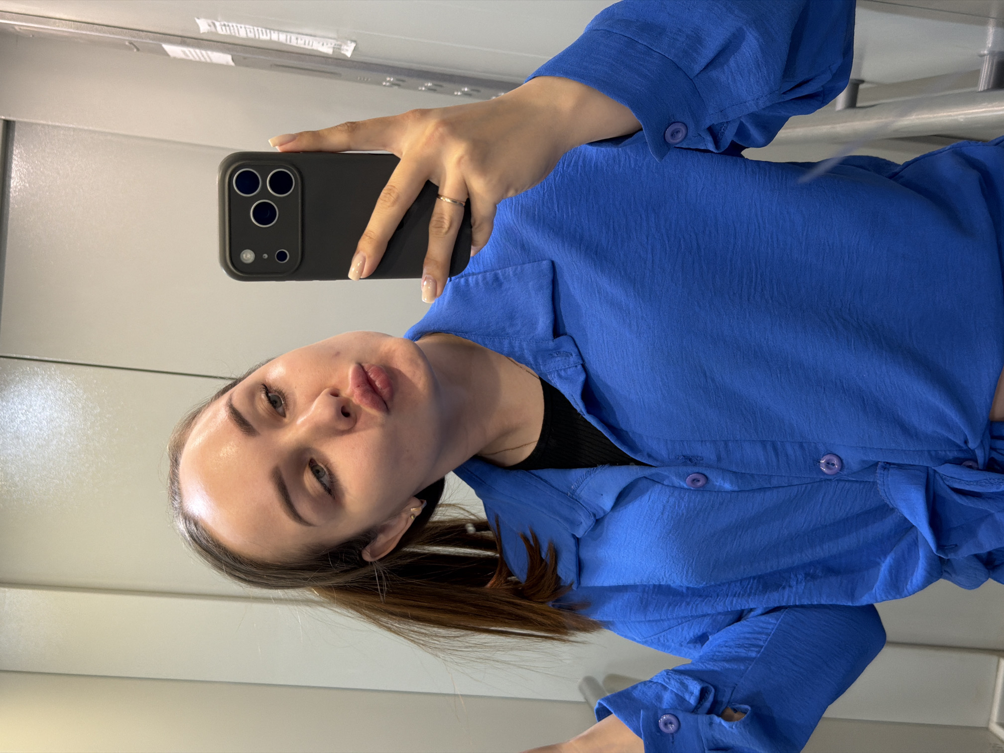 A woman wearing a blue button-down shirt is taking a selfie with a black smartphone. The phone has three lenses. She is holding the phone with her right hand, which has a gold ring on the ring finger. The background appears to be a white wall or door with horizontal lines. The photo was taken in a mirror. There is no evidence in the image that places it in the city of Chaikovsky, Russia.