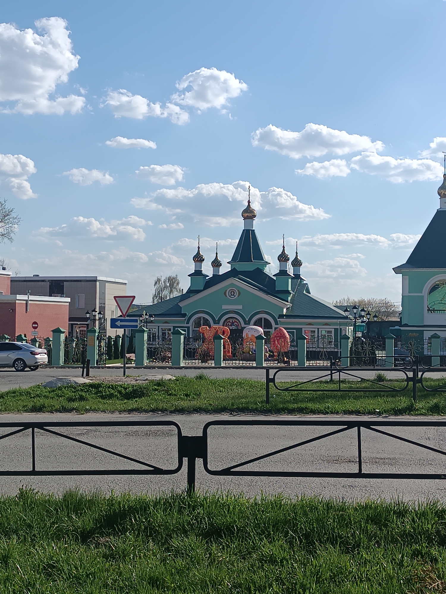 The image shows a green Eastern Orthodox church located in the city of Chaikovsky, Russia. The church features multiple domes with golden onion-shaped tops and is surrounded by a decorative green metal fence. Large, ornamental Easter egg decorations can be seen inside the fence. In the background, other buildings are visible, including a two-story building with a light brick exterior. The sky is blue with scattered white clouds. A road with a black metal safety barrier is in the foreground, with grass growing on either side. A car can be seen driving on the road. A street sign is visible, indicating the direction of traffic.