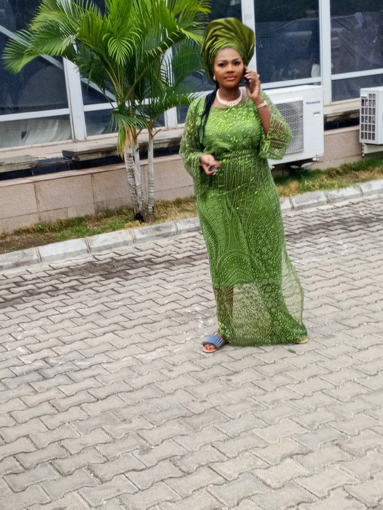 An African woman, appearing to be of medium complexion, stands outdoors, talking on a smartphone held to her right ear. She is dressed in a traditional emerald green long gown featuring intricate patterns and possibly sequins or embroidery. A matching green gele (headwrap) adorns her head. She wears a gold-colored multi-strand necklace and a gold-colored watch on her left wrist. On her feet are sandals with blue and reddish-orange straps. Her left hand is slightly raised, holding a small dark object.
The setting appears to be a paved outdoor area, possibly a walkway or parking lot, made of grey interlocking paver stones. Behind her is a light-colored building with large dark windows and white frames. To the left of the woman (viewer's left) is a small palm tree with vibrant green fronds. To her right (viewer's right), two white outdoor air conditioning units are mounted on the building wall; the larger unit prominently displays the brand name "Panasonic." A concrete curb separates the paved area from a narrow strip of dry grass and dirt next to the building. The scene takes place during the daytime, likely under bright or overcast conditions as no harsh shadows are visible. The overall context suggests a location in Barakin Lerawi, Nigeria.