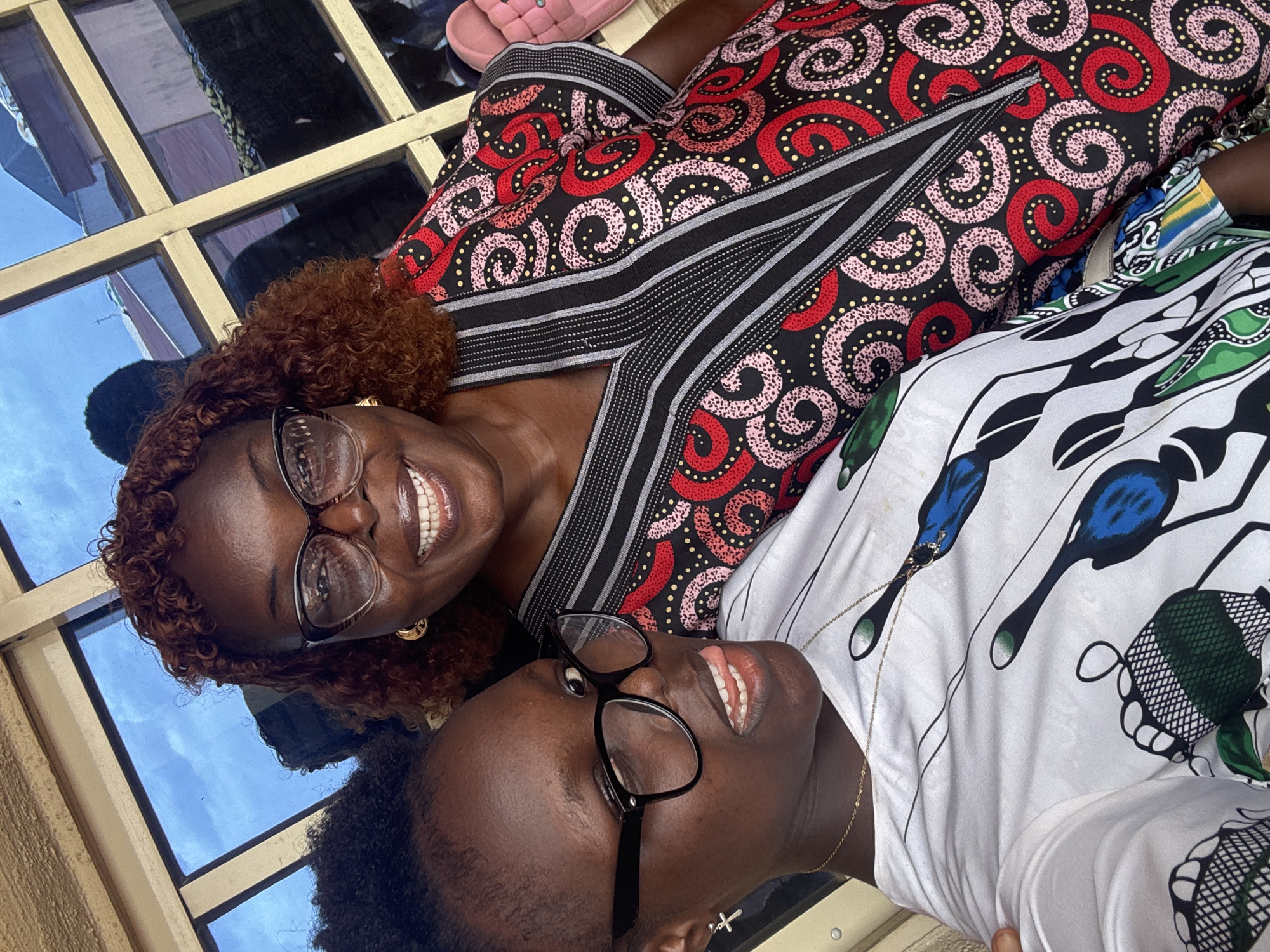 Two Black women are smiling at the camera in a close-up shot. The woman on the left has brown curly hair and is wearing glasses and a dress with a red and black pattern. The woman on the right has short black hair and is wearing glasses and a white shirt with a blue and green design. They are indoors, near a window. The location is Onitsha, Nigeria.