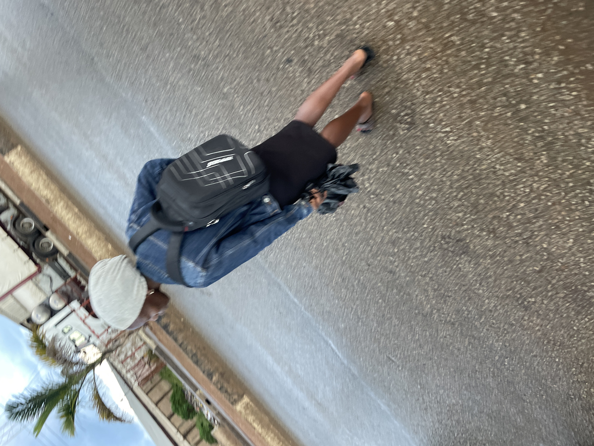 A young Black female wearing a light-colored headscarf, a denim jacket, and a black skirt is walking on a wet asphalt surface. She carries a black backpack with white accents and a folded black umbrella in her left hand. The scene appears to be outdoors on a street, with a portion of a white truck and a palm tree visible in the background. The location is identified as Barakin Lerawi, Nigeria, and the wet surface suggests recent rainfall. The image is taken from a slightly elevated, rear-facing perspective, with motion blur indicating movement.