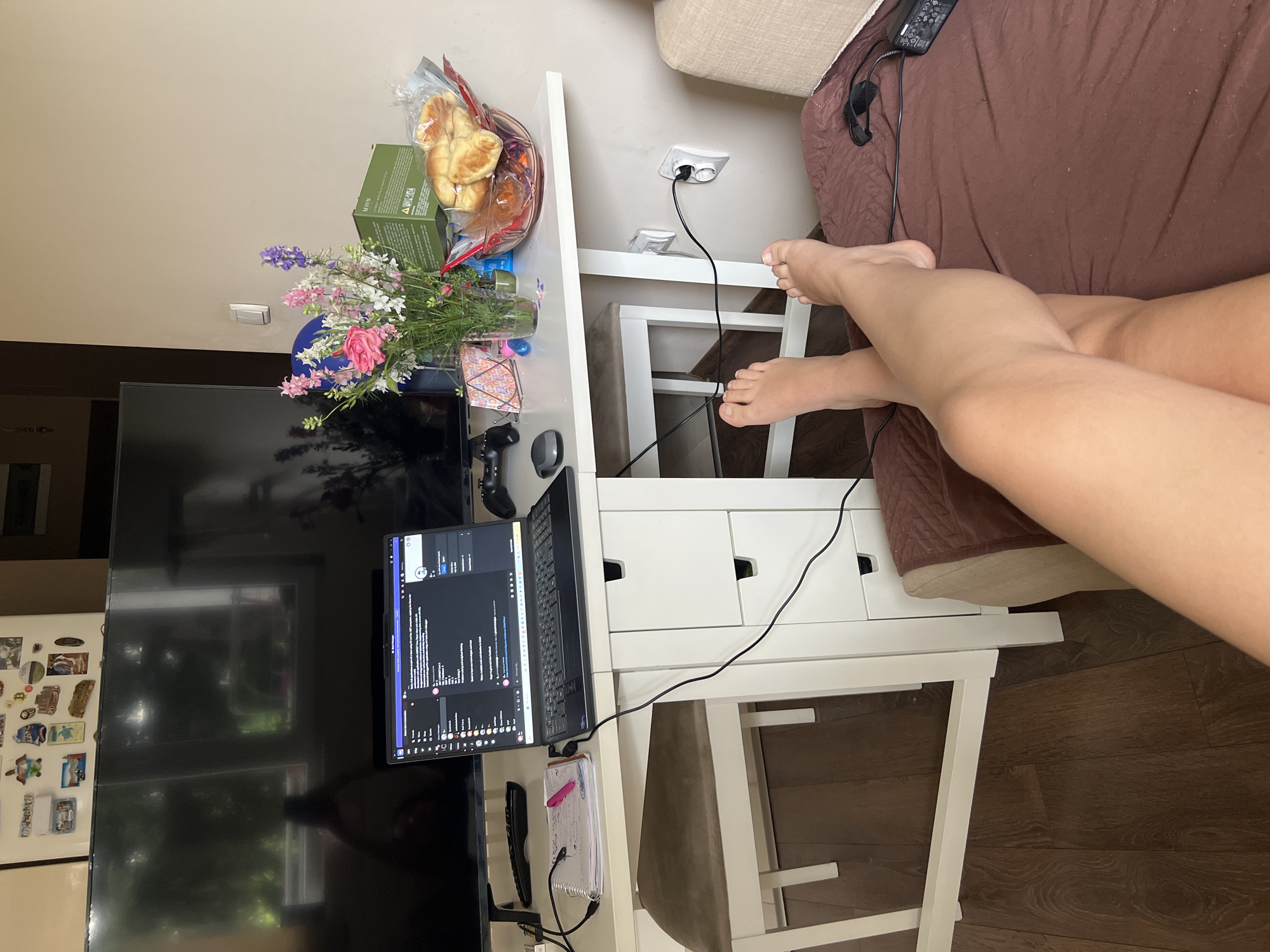 A top-down shot captures a domestic scene with a focus on a small white desk and a person's legs. The desk is laden with various items. On the left, a large black television screen dominates, with a laptop open to a screen displaying code or text in front of it. Next to the laptop is a notebook with a pink pen, and a black remote control. To the right of the laptop, on the desk, is a bouquet of colorful flowers in a glass jar, and a plastic container filled with pastries. The desk also has drawers, and a black cable runs from an electrical outlet on the wall to the laptop.
The lower portion of the image shows the bare legs and feet of a person, positioned crossed at the ankles, resting near the desk. The person is wearing dark-colored headphones around their neck, with the wires also visible. The background suggests a home environment with a brown sofa and wooden flooring. The overall lighting implies it's daytime.
The presence of the laptop with code, the notebook, and the remote control suggests the person might be working, studying, or enjoying leisure time at home. The pastries and flowers add a touch of domesticity and comfort to the scene. The location context provided is Sofia, Bulgaria, but this is not directly discernible from the image itself.