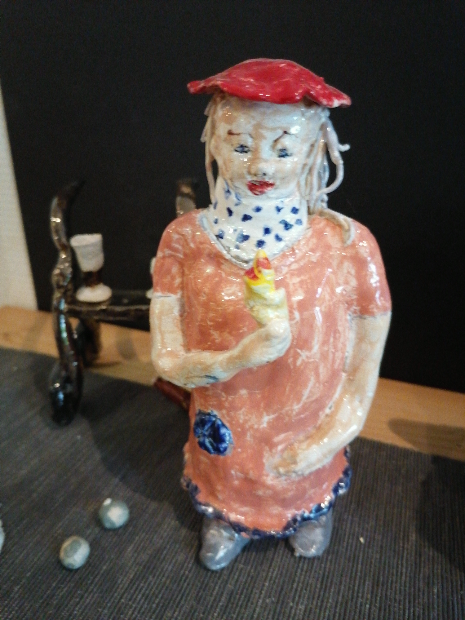 This is a forensic, matter-of-fact description of an image. The scene shows a ceramic figurine of a person wearing a red hat, a blue and white polka dot scarf, and an orange dress with a blue flower on the skirt. The figurine is holding a yellow and red object in its right hand. The figurine is standing on a dark gray, ribbed surface, possibly a rug or mat. Behind the figurine, on the left, is a dark, curved object, possibly part of a stand or another decorative item. In the background, the wall is black. There are two small, round, gray objects on the surface in front of the figurine, possibly decorative stones or marbles. The image does not provide any information about the location in the city of Grünwald, Germany.