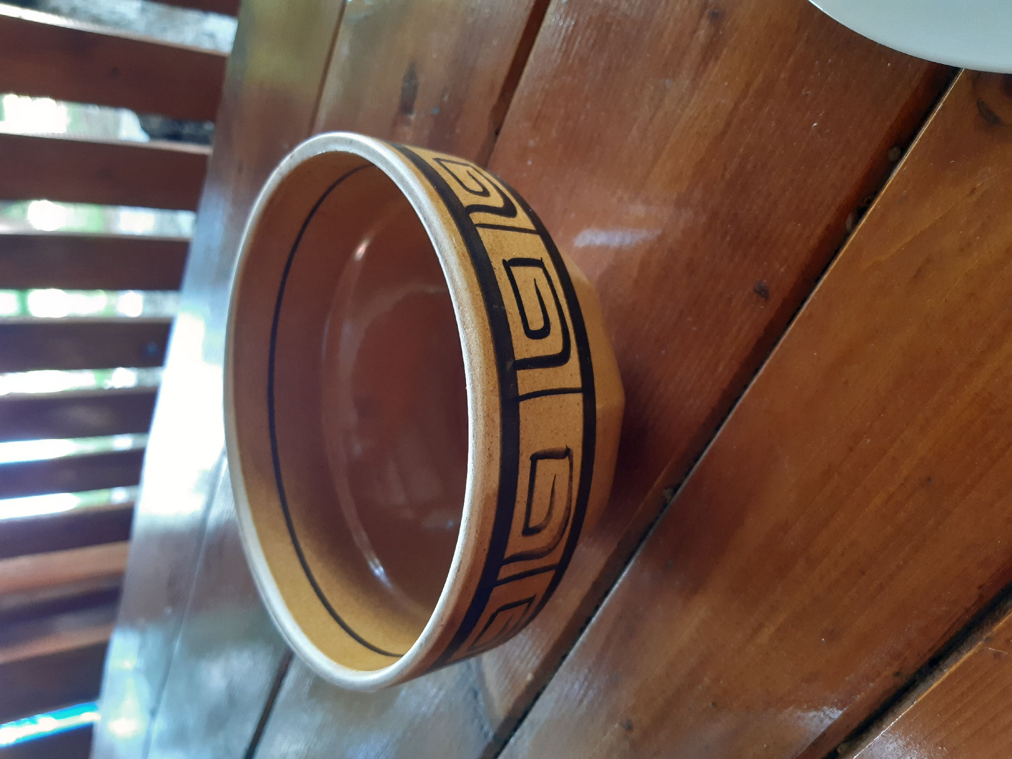This media file features a close-up, angled shot of a ceramic bowl with a decorative geometric pattern. The bowl is a warm, earthy orange-brown color on the inside and has a band of black, maze-like designs around the rim. A thin black line encircles the exterior of the bowl just below the rim. The bowl rests on a wooden surface, likely a table, composed of several planks with a visible grain and a glossy finish. The lighting suggests it is daytime, with bright light reflecting off the wooden surface and the bowl.
In the background, to the left of the bowl, is a structure made of horizontal wooden slats, partially obscuring the view beyond. Through the gaps in these slats, blurred greenery and bright light can be seen, hinting at an outdoor setting, possibly a porch, patio, or a sheltered area with natural light. The overall impression is one of a rustic and natural environment, consistent with a location like Kavetchyna, Ukraine, which is known for its rural landscapes. No people are visible, and the focus is entirely on the bowl and its immediate surroundings. The image does not contain any visible text.