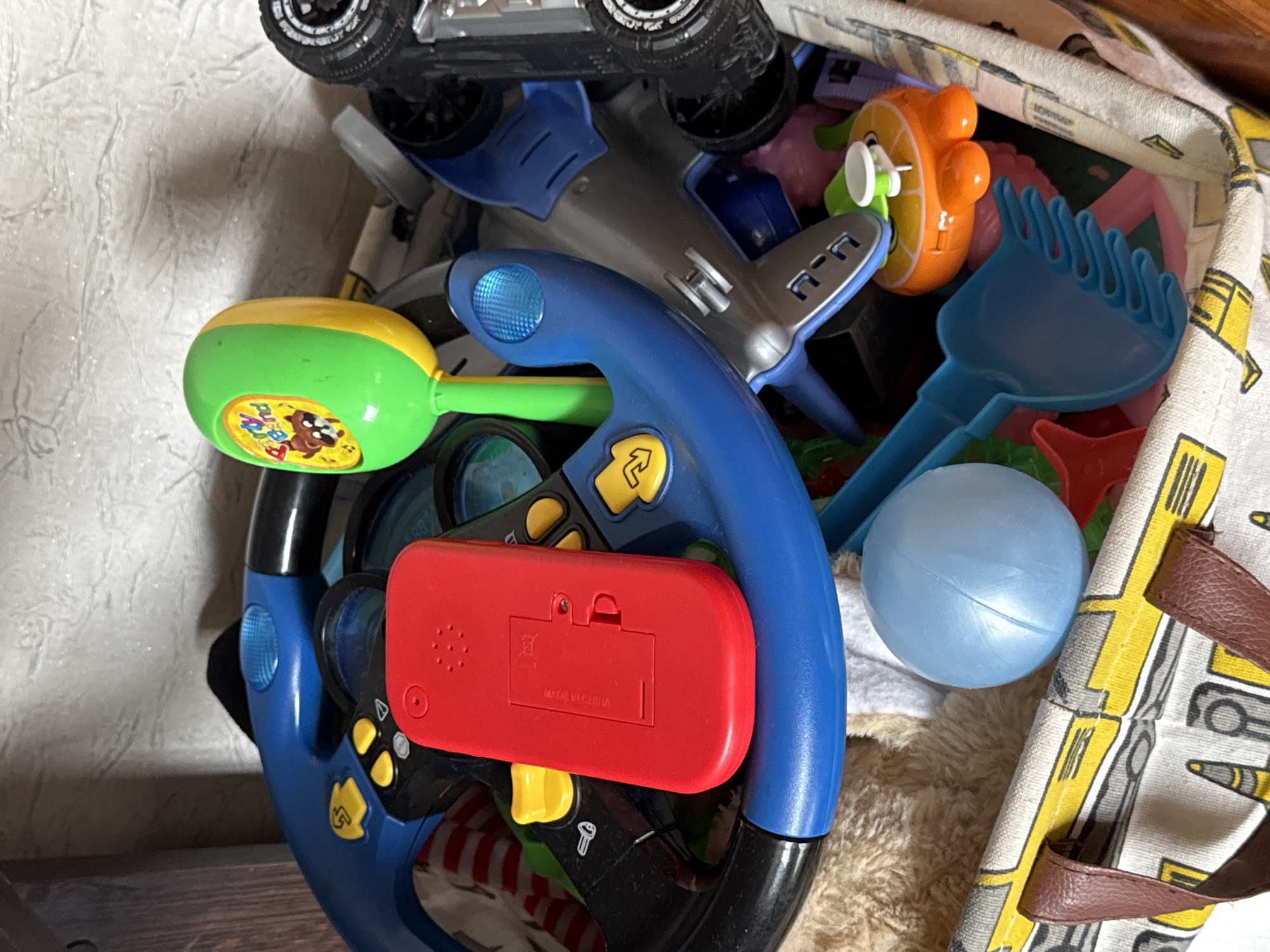 The image shows a close-up, overhead view of a toy bin filled with various children's toys. The toys are colorful and appear to be made of plastic. Visible objects include a toy steering wheel, a toy airplane, a blue plastic rake, a light blue plastic ball, and a toy resembling an orange slice with a white button. There is also a red battery compartment with "MADE IN CHINA" text printed on it, as well as a battery symbol. The bin itself is decorated with a pattern featuring yellow and gray geometric shapes, possibly resembling vehicles. The overall scene is a cluttered collection of toys, suggesting a child's play area or storage space. The lighting appears to be indoor, and there are no indications of time of day or weather. The location context provided is "Яромінскі сельскі Савет, Belarus," which is a rural council in Belarus.