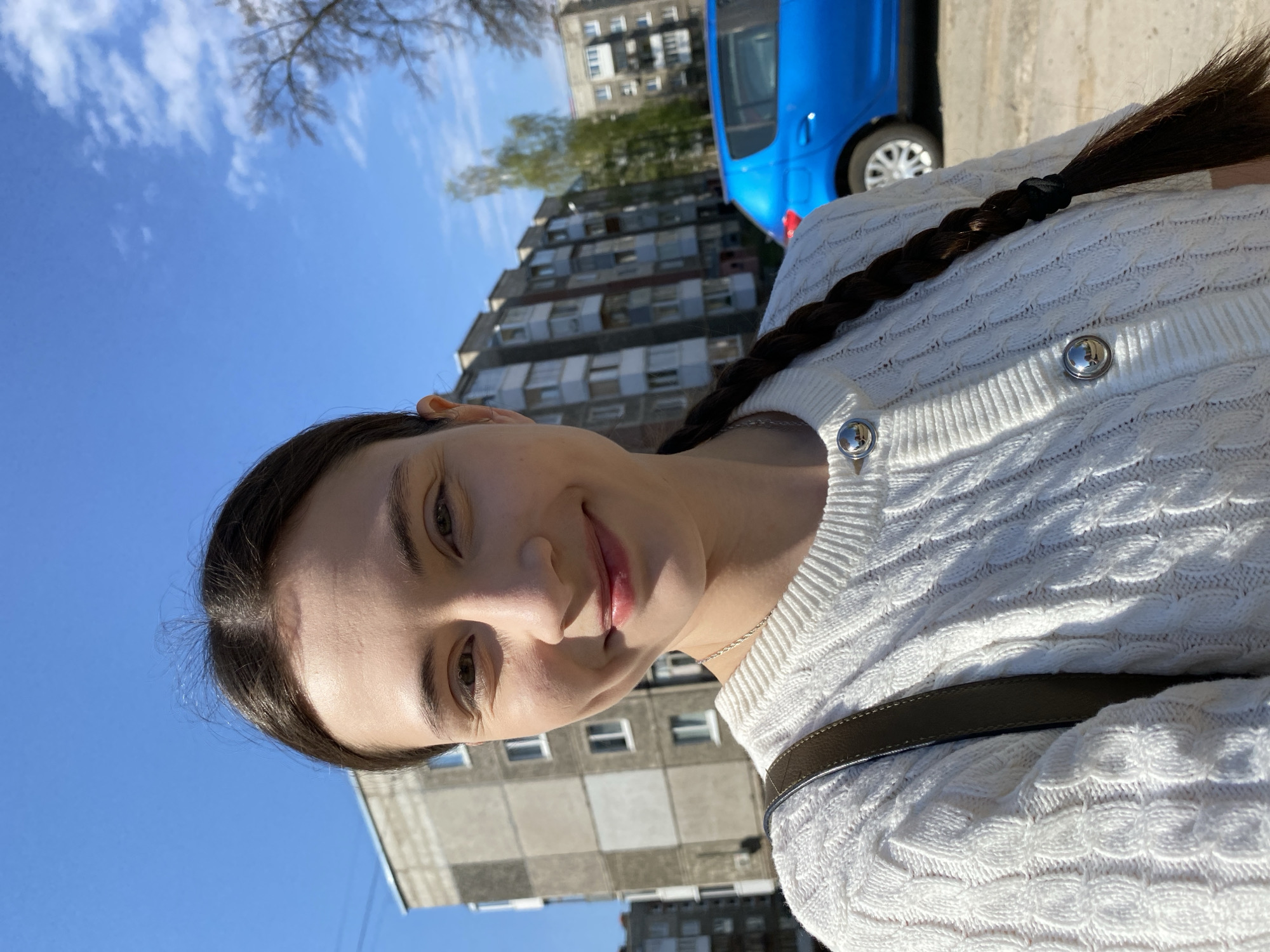 A woman is pictured in a medium close-up shot, angled slightly upward. She has fair skin, dark hair in a braid, and wears a white sweater with silver buttons. A brown bag strap is visible across her chest. She has a slight smile. Behind her is a clear blue sky with a few clouds and the top of a bare tree. A multi-story apartment building is in the background, as well as a blue car. The photo appears to be taken outdoors during daylight hours. The location is Votkinsk, Russia, identifiable by the style of the apartment building and the presence of a Lada car, both common in the region.