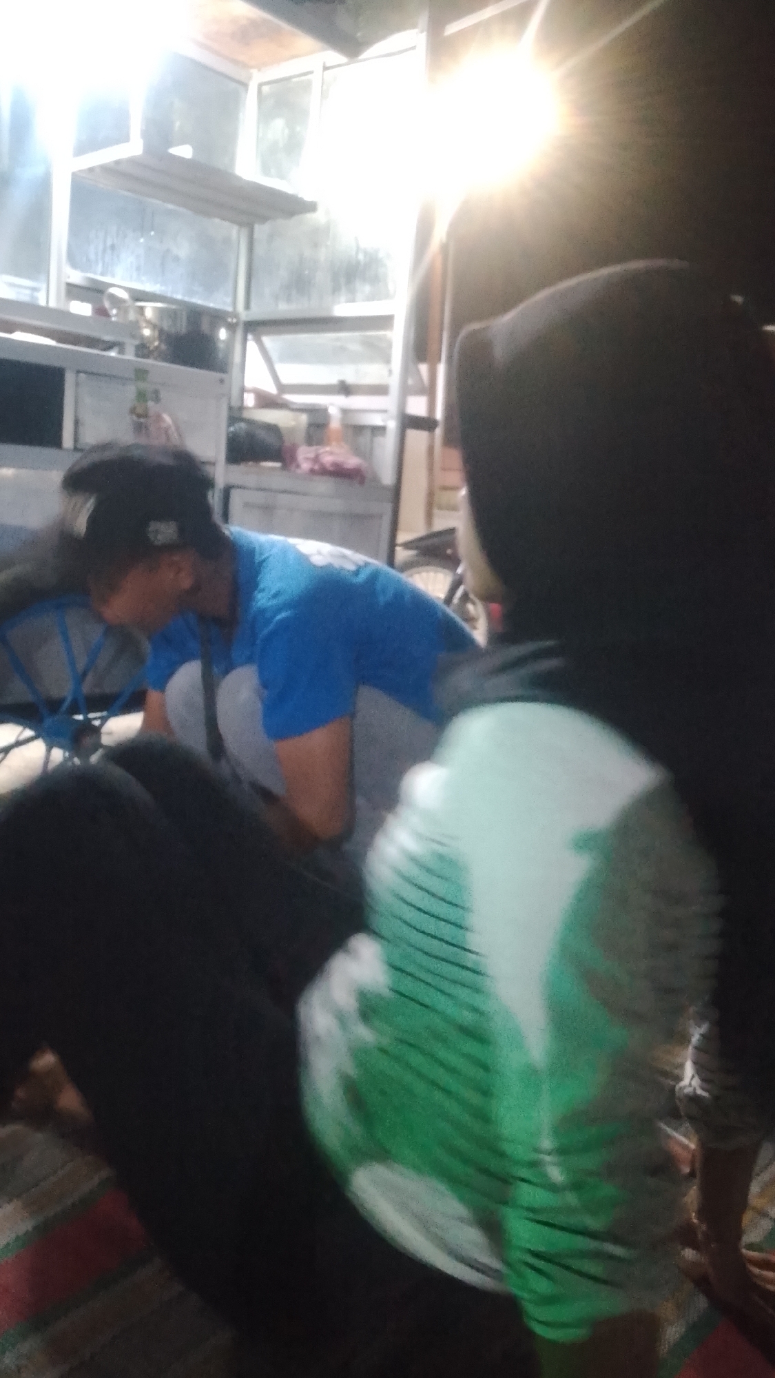 In Karanglangu, Indonesia, a dimly lit interior scene features two individuals. In the foreground and slightly right, a person wearing a black hijab and a green and white patterned top is partially visible, facing away from the camera. To the left and further back, a person wearing a blue t-shirt and a black baseball cap is squatting down, looking downwards. The environment appears to be a stall or shop with shelving units and a bright light source casting a glare on the upper right portion of the image. Objects on the shelves are indistinct. The floor is covered with a patterned rug.