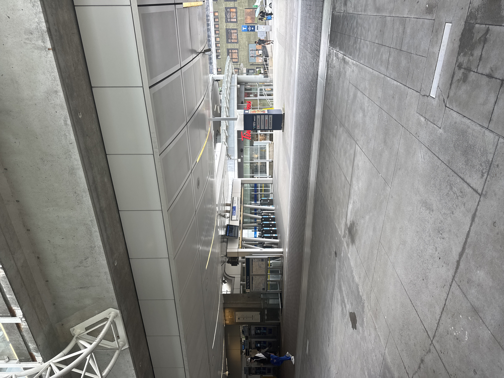 The image depicts an outdoor scene at a transportation hub in New Westminster, Canada. In the foreground, a paved area with visible seams and some damp patches is shown. The midground reveals the entrance to a transit station, characterized by ticket gates, information displays, and a prominent sign for "Tim Hortons" with a list of other businesses. A blue "T" logo, indicative of TransLink, is visible on a pole to the right. Several individuals are present: one person is walking away from the camera on the left, a couple of people are walking towards the right in the distance, and another individual is walking in the background to the right of the Tim Hortons. The background consists of buildings with visible windows and brickwork. Overhead, a concrete structure with white architectural elements is present. The overall environment appears to be urban and functional, designed for public transit and commercial activity.