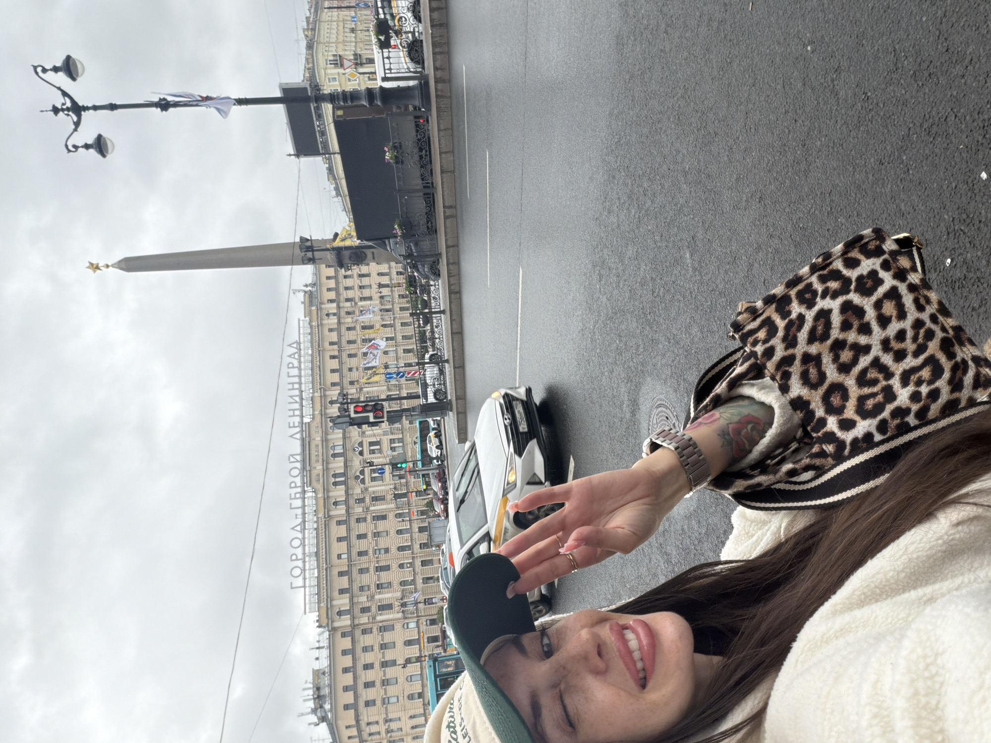 In Saint Petersburg, Russia, a woman with long dark hair and a light-colored fleece jacket takes a selfie outdoors on a cloudy day. She is wearing a beige baseball cap with green trim and a black brim. Her left eye is closed in a wink, and her right hand is raised, obscuring part of her face, with fingernails painted pink. She is wearing two rings on her right hand. A leopard-print bag with black and white striped straps is visible over her right shoulder. Her left arm, adorned with a tattoo of a rose, is also visible.
The background depicts a wide, wet street with cars driving on it, suggesting recent rain. A white Hyundai Elantra taxi with yellow accents is prominently featured. In the distance, a large building with the inscription "ГОРОД-ГЕРОЙ ЛЕНИНГРАД" (City-Hero Leningrad) is visible, along with a tall obelisk topped with a star. A street lamp with three globes stands to the right of the obelisk. Traffic lights are also visible. The overall scene is a street view in Saint Petersburg, likely near a significant monument or historical site.