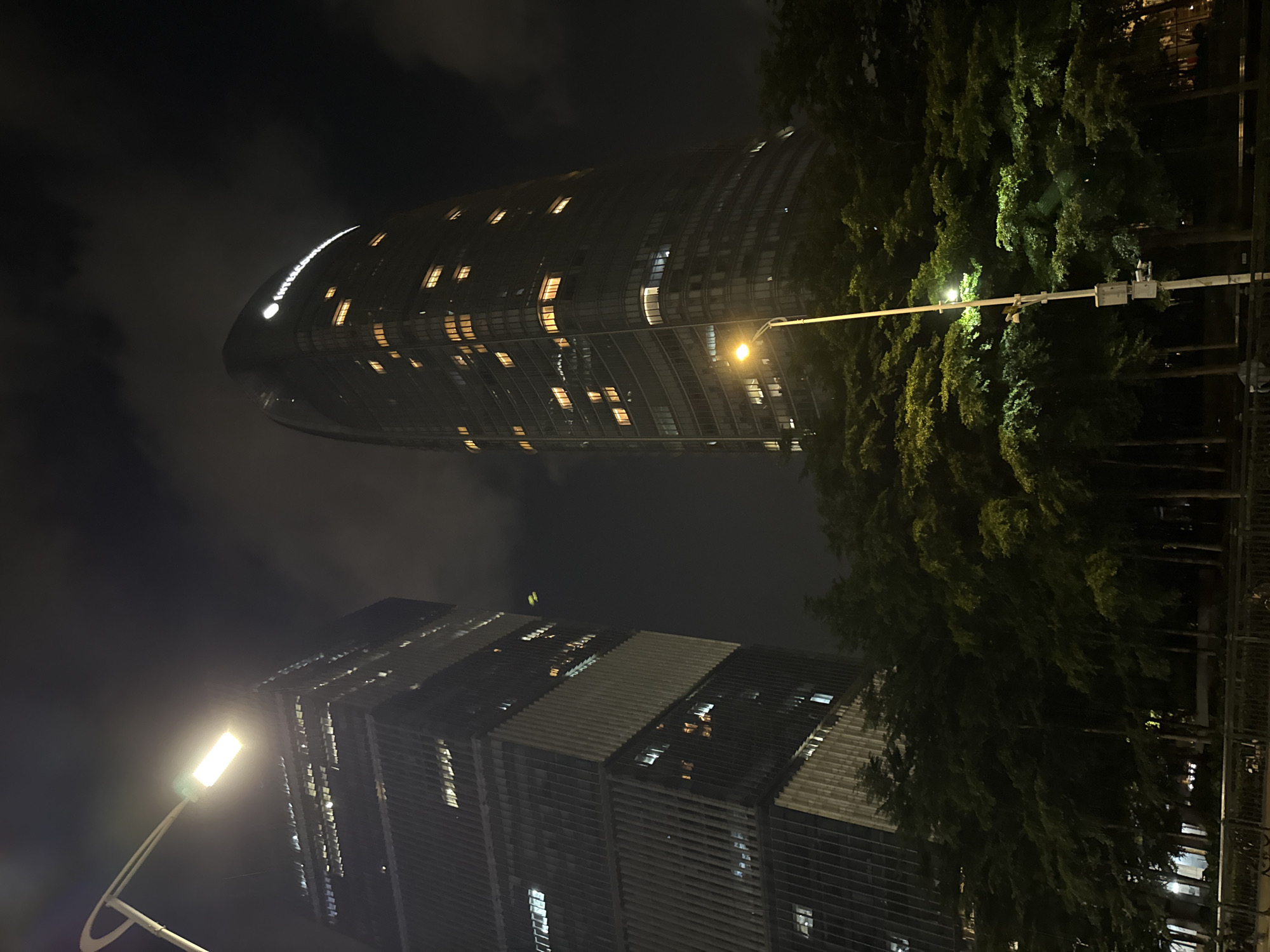 The image captures a nighttime urban scene in Haizhu District, China. Two modern skyscrapers dominate the frame, their illuminated windows creating a geometric pattern against the dark sky. The building on the left is uniquely curved, resembling a crescent moon or a gentle wave, and its upper section is crowned with a bright, illuminated logo or text that is not clearly legible. The other building, to the right and lower in the frame, is a more traditional rectangular skyscraper with a facade featuring horizontal louvers or panels, also dotted with lit windows.
A streetlamp with a bright white light is prominently positioned in the foreground, casting a glow on the surrounding elements. Behind and between the skyscrapers, a dense cluster of lush green trees forms a natural barrier, their foliage illuminated by the streetlamp and the building lights, creating a striking contrast between nature and the urban environment. The sky is dark and cloudy, suggesting a typical night in the city. The overall mood is one of quiet nighttime activity, showcasing the architectural modernity of the city juxtaposed with natural elements. No people are visible in the frame, and the focus is solely on the buildings, trees, and streetscape. The image appears to be taken at an angle, giving a dynamic perspective of the urban landscape.