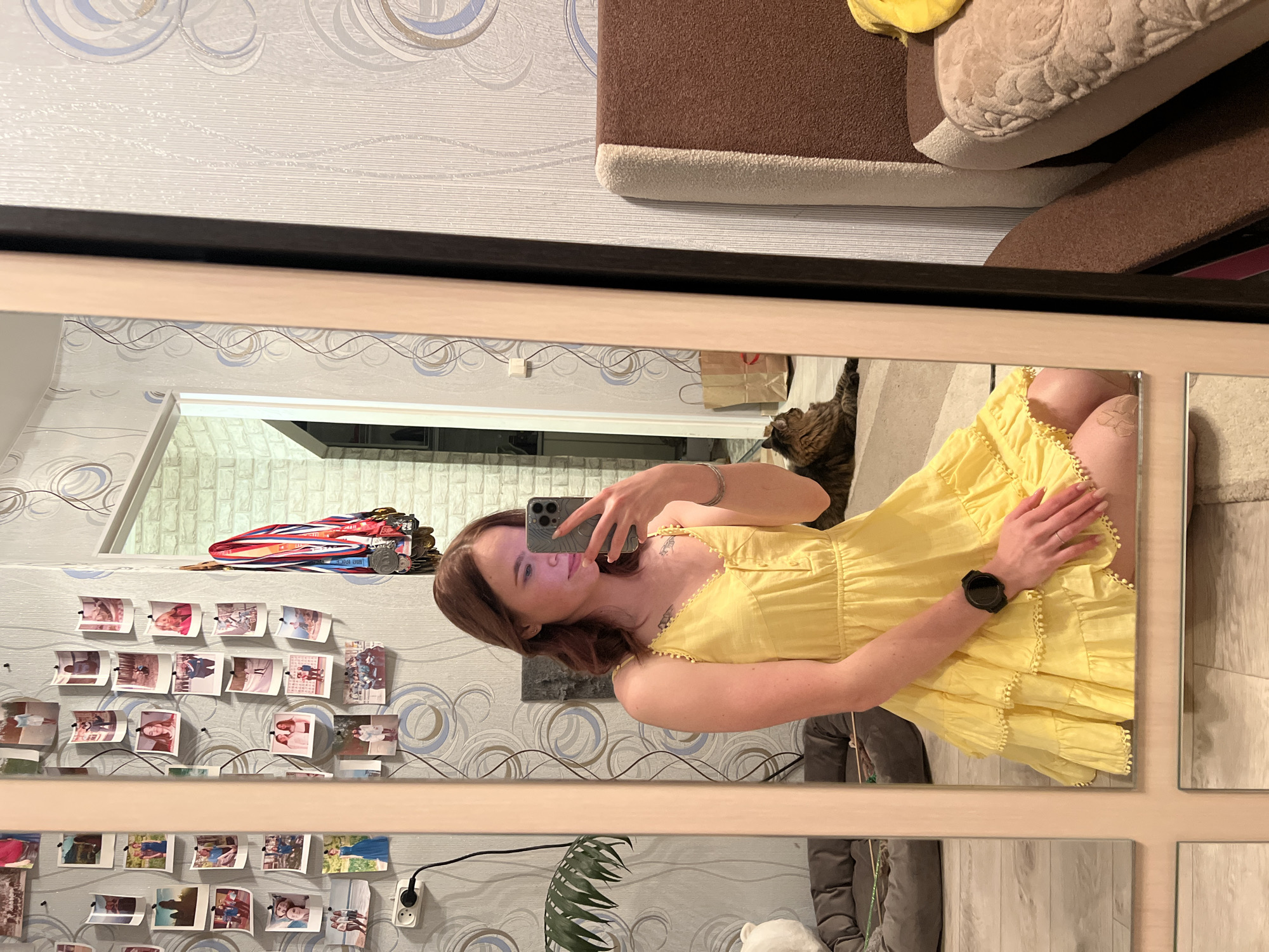 A young woman is taking a selfie in a mirror. She is wearing a yellow dress and a black watch. The mirror reflects a room with wallpaper, photos, medals, a plant, and a couch. The photo may have been taken in Chaikovsky, Russia.