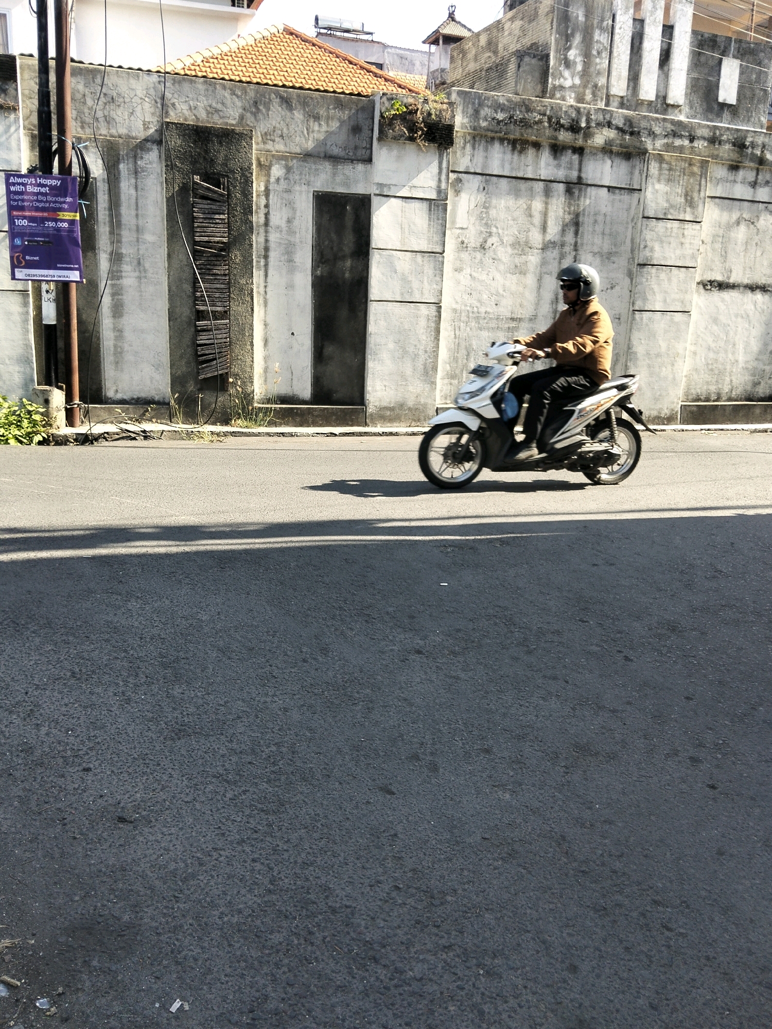 In Kuta, Indonesia, a person wearing a helmet and sunglasses rides a white scooter down a paved road. The rider is dressed in a brown jacket and black pants. The scooter has black and silver detailing. The rider is positioned in the middle right of the frame, moving from left to right. Behind the scooter, a textured concrete wall runs horizontally across the image, featuring vertical paneling and some discolored sections. To the left of the wall, a black utility pole stands, with a purple advertisement attached to it. The road surface is asphalt. Sunlight casts shadows across the road. The environment appears to be an urban setting with a building visible in the background.