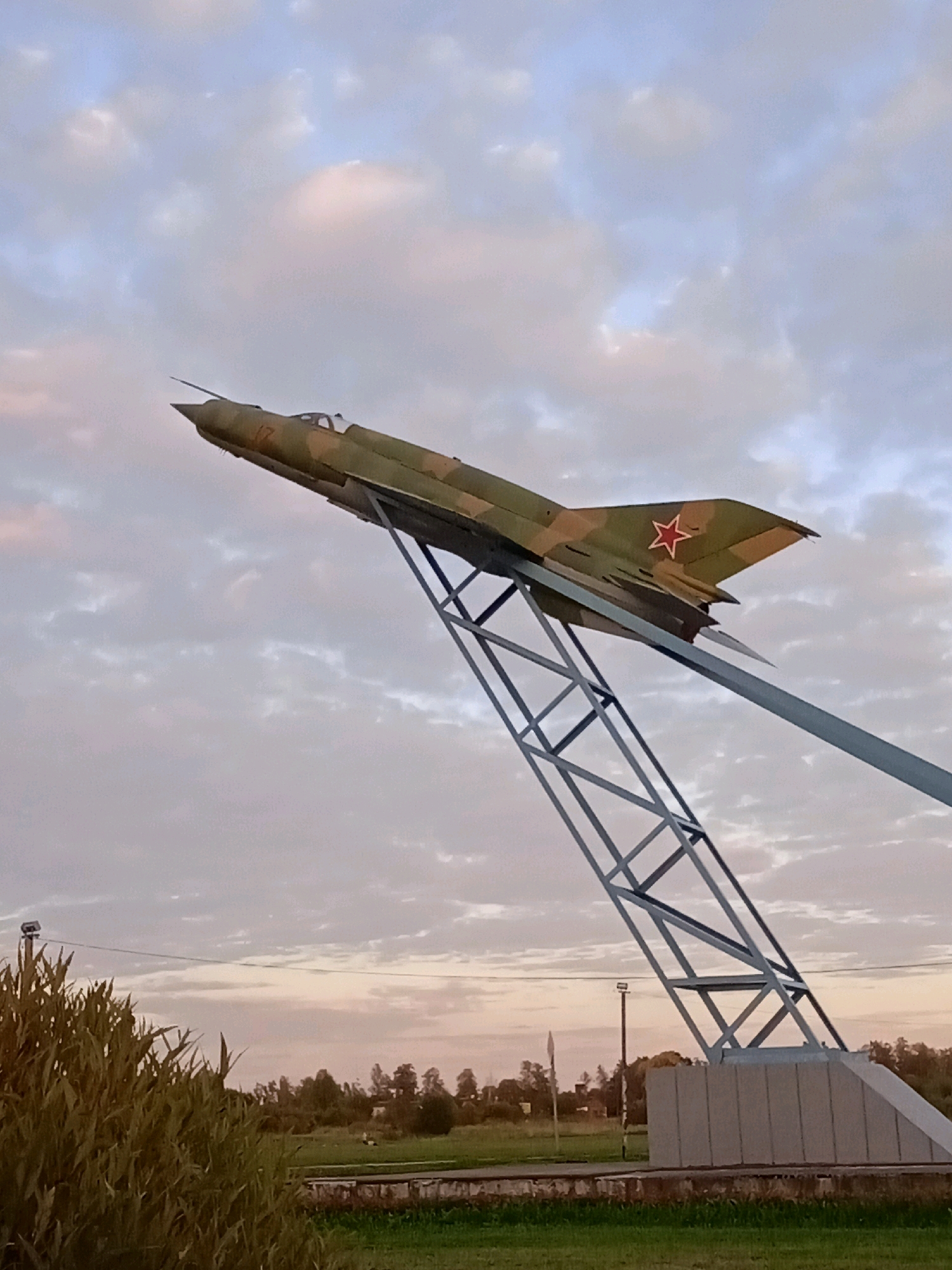 An outdoor, eye-level, full shot shows a camouflage-painted fighter jet monument suspended on a metal support structure. The jet is angled upwards and to the right. It is painted in a pattern of green and tan camouflage, with a red star on its tail fin. The support structure is a gray, open lattice of metal beams. The jet is positioned against a pale blue sky with scattered white and pinkish clouds. In the foreground, there is tall, green grass and some darker foliage on the left side of the frame. In the background, there is a flat, green field with scattered trees and some buildings in the distance. A concrete base supports the metal structure. A utility pole with a light is visible in the background.