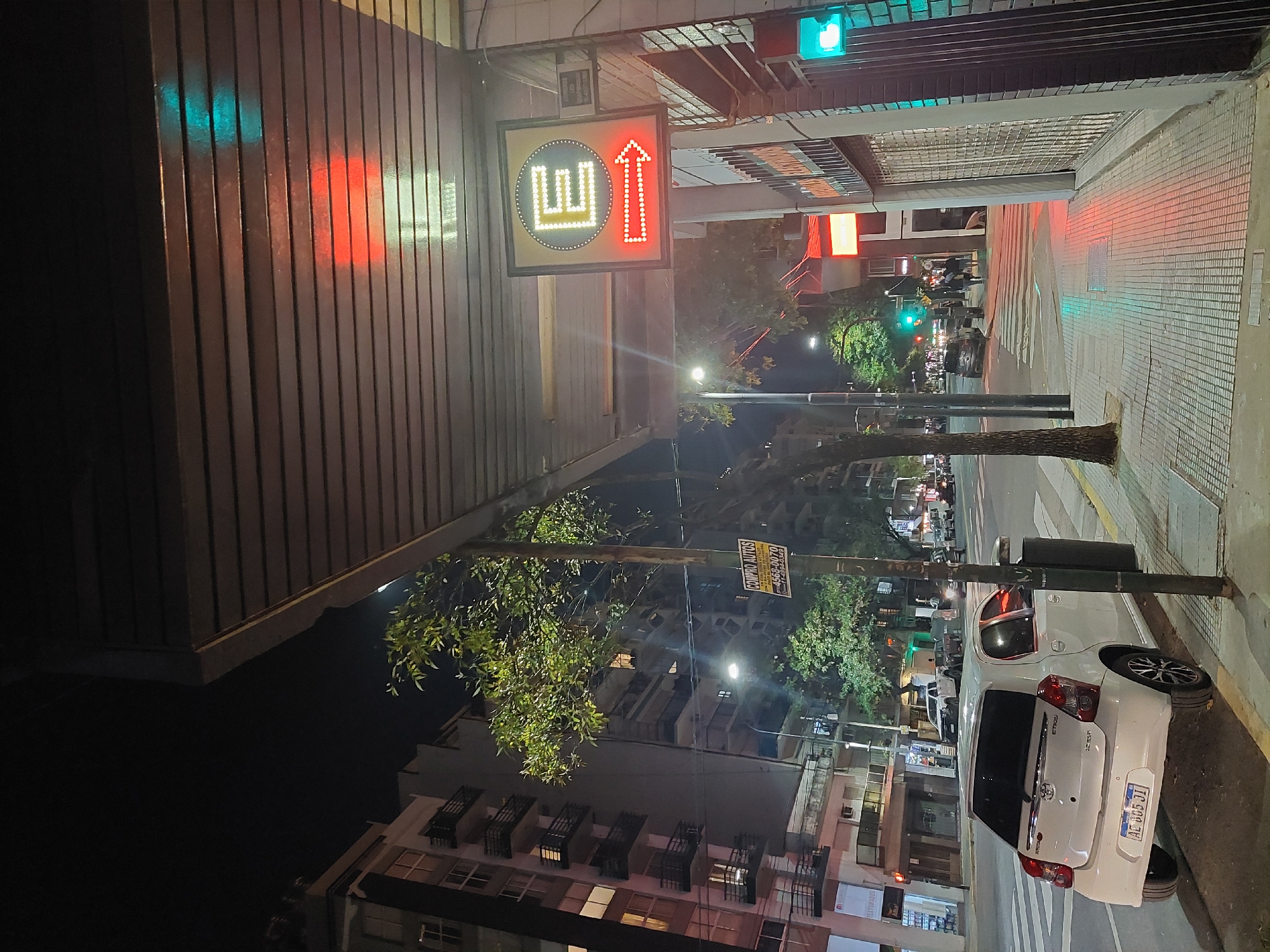 This is a nighttime photograph of an urban street scene. The perspective looks down a street lined with buildings and trees, illuminated by artificial light.
On the left side of the frame, a building with vertical slatted siding reflects green and red light from nearby traffic signals. Hanging from an awning of this building is a square, illuminated sign featuring a stylized yellow "W" symbol on the left and a red upward-pointing arrow on the right, both composed of individual lights, set within a black border with yellow dots. Above this sign, a green traffic light is visible.
The street extends into the distance, with sidewalks on both sides. On the right side of the street, a white Toyota Etios car is parked at the curb, its rear facing the viewer. The car's license plate reads "AE 805 JI". A tree trunk stands nearby. Further down the street, more buildings are visible, some with balconies and illuminated windows. Overhead, another green traffic light is prominent.
Amidst the foliage of trees in the middle distance, a yellow banner is partially visible, displaying the Spanish text "COMPRO AUTOS" (I buy cars) and a phone number "4568-0070". Several streetlights cast bright glows, and distant figures can be seen walking on the sidewalk. The general ambiance is that of a bustling city evening. The visible Spanish text and the license plate corroborate the provided context of Buenos Aires, Argentina.