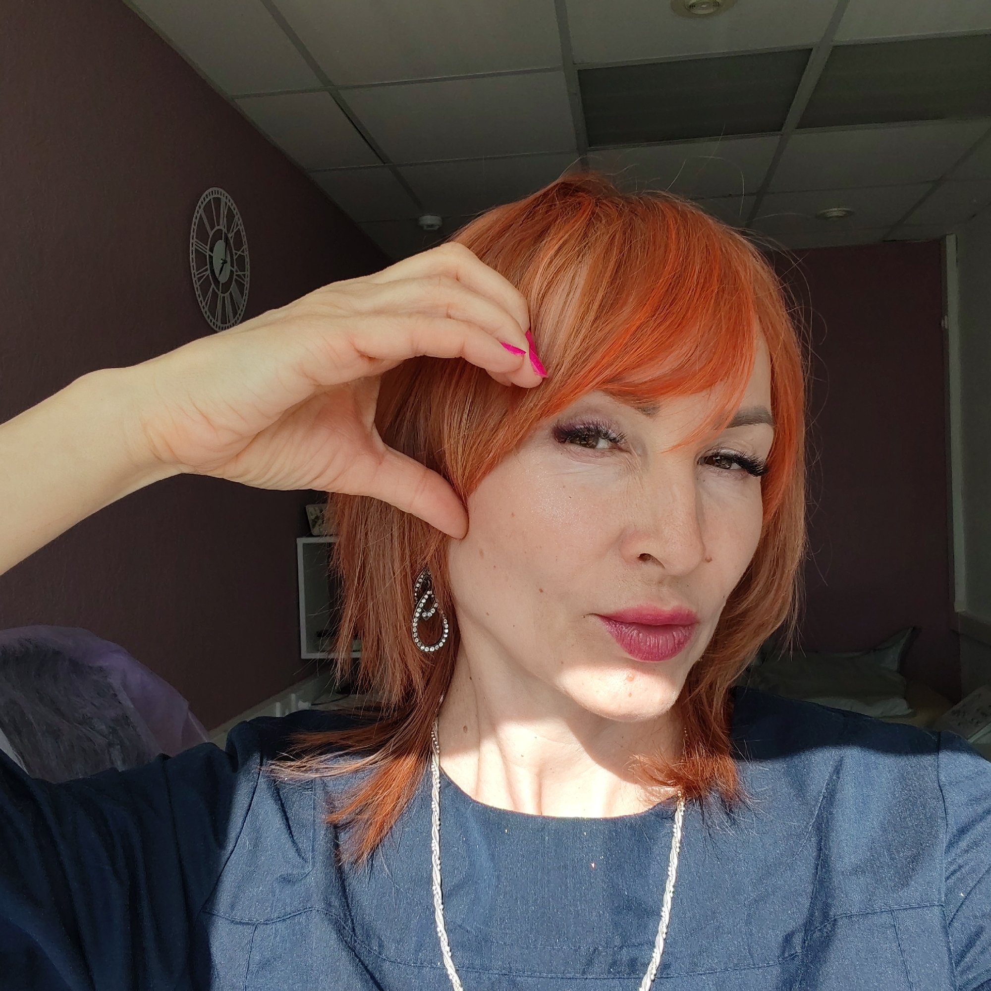 A woman with vibrant orange hair, pink nails, and maroon lipstick poses for a selfie indoors. She wears a dark blue shirt and a silver necklace. Her left hand is raised, with her fingers touching her forehead and cheek. She is wearing a pair of dangle earrings. The background includes a purple wall with a decorative clock, a white cabinet, and a bed with pillows. The location is potentially in an indoor establishment in the city of Chaikovsky, Russia.