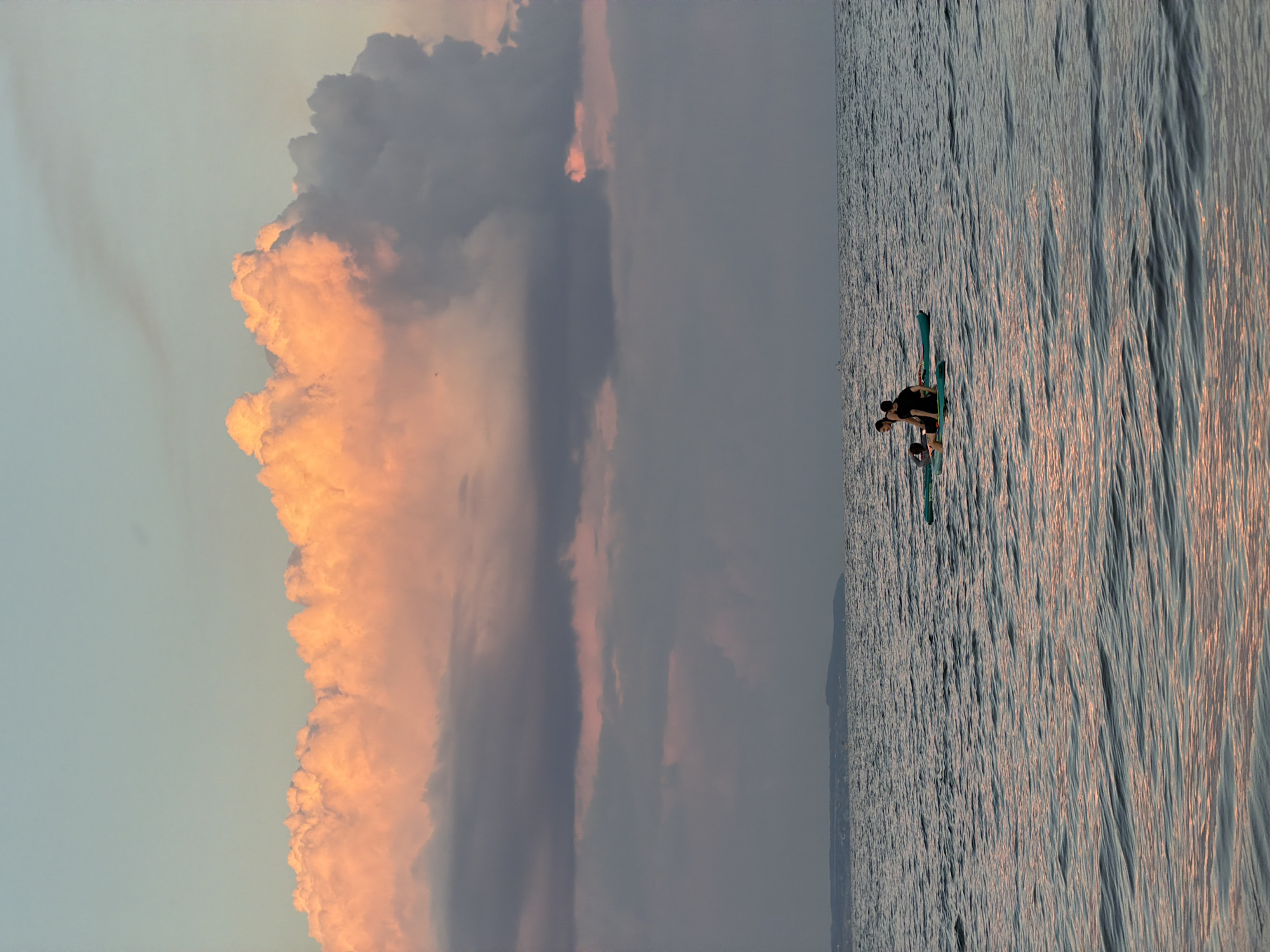 A wide shot captures a serene seascape at sunset, with a hint of a distant shoreline. The sky is a canvas of soft pastel hues, dominated by a large, fluffy cloud illuminated with a warm, peachy glow from the setting sun. Below this vibrant cloud formation, darker, muted clouds stretch across the horizon. The water is a deep blue-grey, reflecting the colors of the sky and creating gentle ripples. In the middle ground, three individuals are present on two paddleboards. One paddleboard has two people, one seated and one leaning forward. The other paddleboard, slightly ahead, has a single person lying on their stomach. The scene is tranquil and evokes a sense of calm as the day draws to a close. The location is identified as the city of Laishevo, Russia.