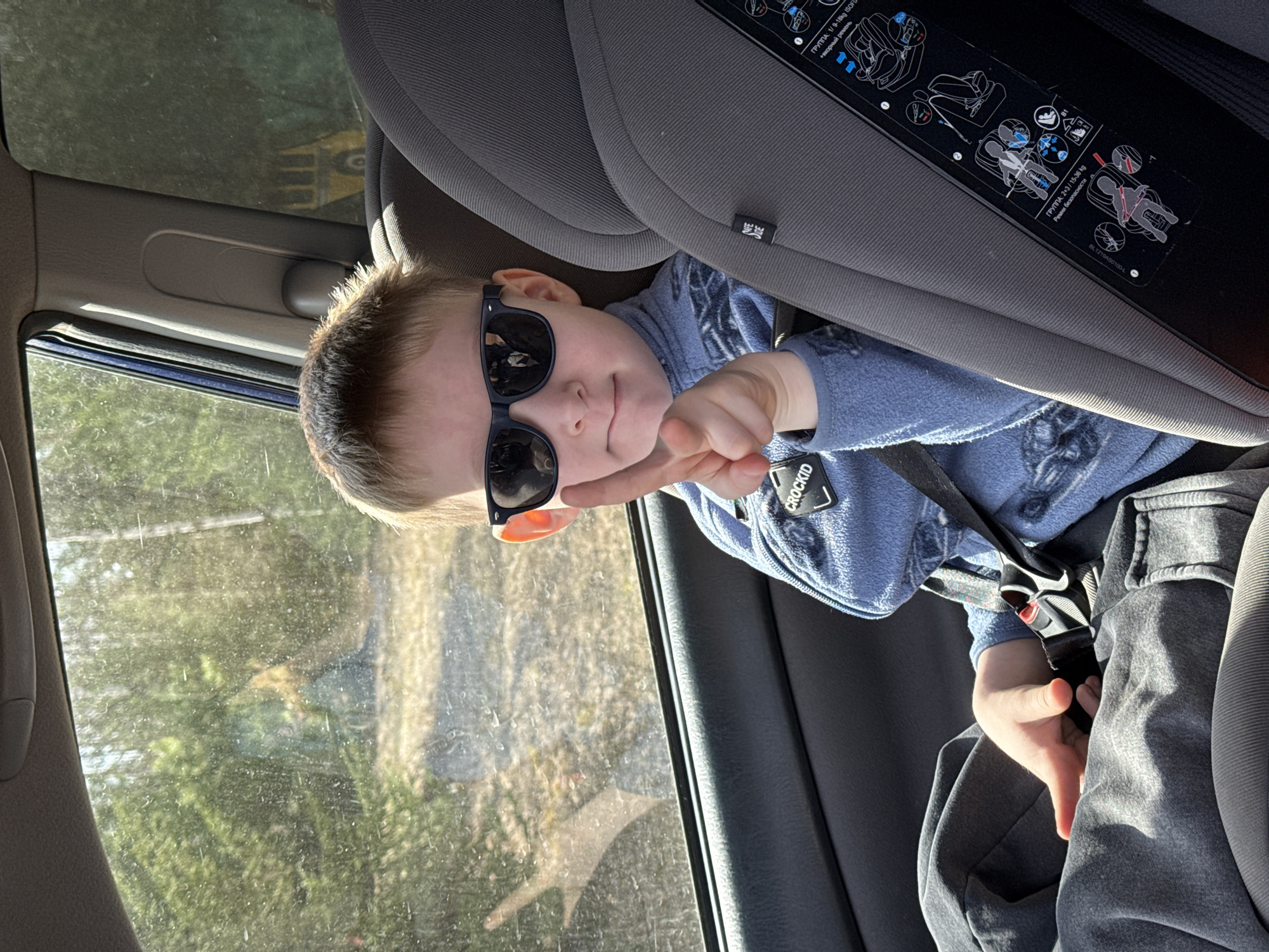A young boy is pictured sitting in a gray car seat inside a vehicle, likely in Малая Уса, Russia. He is wearing dark sunglasses and a blue long-sleeved top, along with gray pants, and is secured by the car seat's harness. The boy looks playfully towards the camera, smirking, and extends his right hand with a "peace" or "victory" sign. Sunlight streams in from the window to his left, indicating a bright, sunny daytime setting. Outside the window, green foliage, trees, and hints of a structure or another vehicle are visible, suggesting an outdoor or rural environment. The car seat features several white safety labels with black text, including "CROCKID", "ГРУППА: 1/ 9-18kg ISOF", "Ремни безопасности", "ГРУППА: 2+3/15-36 kg", "BL1219AS0100-L", and "+якорный ремень", providing instructions for its use with different weight groups and belt types. The overall scene captures a candid, playful moment of a child traveling in a car.