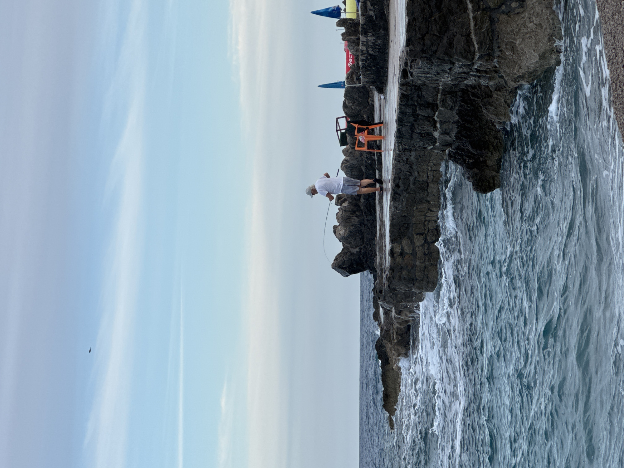 An outdoor, eye-level, full shot captures a person fishing from a rocky pier on the coast of Prijevor, Montenegro. The sky is a soft blue with wispy white clouds, and a faint object, possibly a bird or drone, is visible in the upper left corner. The sea is a deep blue with white-capped waves crashing against the dark, rugged rocks of the pier. A person, wearing a white t-shirt, grey shorts, and a white cap, stands on the pier holding a fishing rod. To the right of the person, an orange plastic chair and a dark metal frame are visible. In the background, two bright blue beach umbrellas and a red object are partially obscured by the pier and rocks. The overall atmosphere is serene and tranquil, with the sound of the waves and the solitude of the fishing activity.