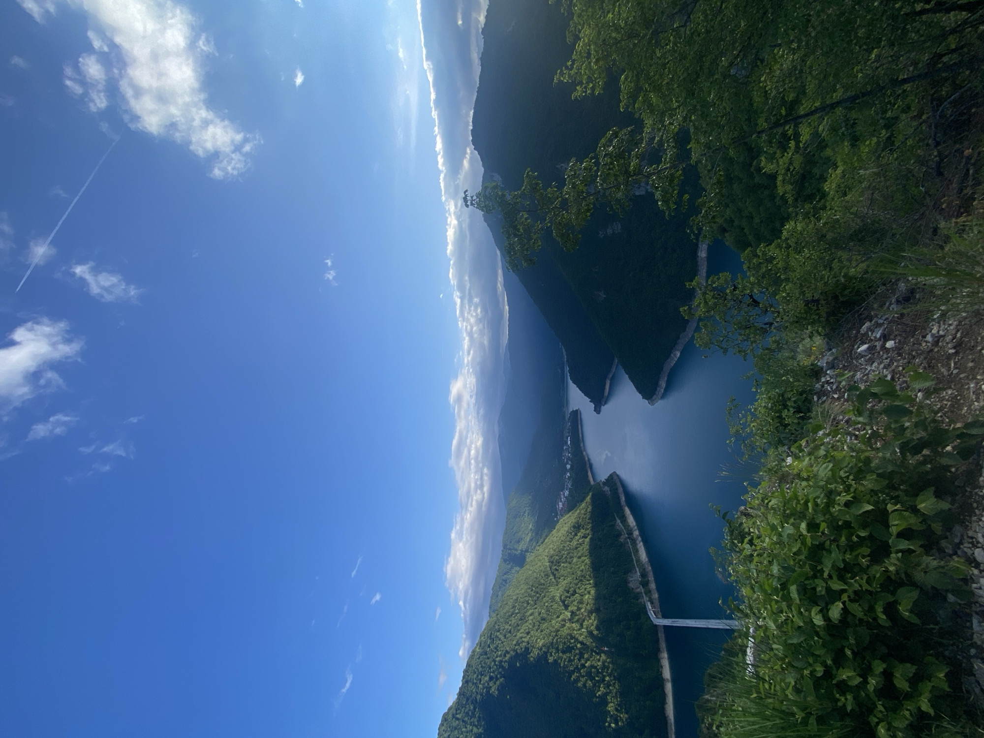 This media file captures a panoramic view of a vast natural landscape in Plužine, Montenegro, likely depicting Piva Lake and its surrounding canyons. The image is taken from an elevated perspective, looking down over the scene.
The dominant feature is a bright blue sky, clear with scattered white cumulus clouds. A thin, white contrail from an airplane is visible in the upper left portion of the sky, stretching diagonally across the frame.
Below the sky, rolling mountains covered in dense, lush green forest dominate the mid-ground and background. The contours of the mountains are rugged, with some areas appearing darker due to shadows or distance, indicating varied terrain.
Winding through these mountains is a deep blue, almost turquoise, body of water – a lake or a wide river. Its serpentine shape carves through the landscape, with several peninsulas and islands formed by the surrounding terrain. In the lower right of the frame, a long, white bridge spans a narrower section of this water body, connecting two landmasses.
In the immediate foreground, dense green foliage, including trees and bushes, frames the bottom and right edges of the image, suggesting the viewpoint is from a vegetated overlook or cliffside. Small stones and earth are also visible on the ground level in the foreground.
The overall impression is one of natural grandeur and tranquility. The scene is brightly lit by daylight, indicating a clear, sunny day, likely morning or afternoon given the illumination. There are no people or visible text in the image.